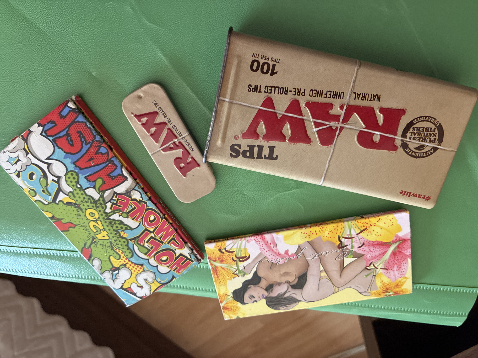 A flat lay photograph displays various smoking accessories arranged on a textured, bright green surface, likely indoors in Varna, Bulgaria.
The central items include a rectangular, light brown cardboard box, branded "RAW TIPS" in prominent red lettering. Text on the box indicates "100 TIPS PER TIN" and "NATURAL UNREFINED PRE-ROLLED TIPS." A thin white string is wrapped around it. The box also states "#rawlife" and features an "AUTHENTIC PUREST NATURAL FIBERS UNREFINED" seal. Adjacent to the box is a smaller, oblong cardboard piece, also bearing the "RAW" logo and "NATURAL UNREFINED PRE-ROLLED TIPS."
To the left, a colorful packet with a comic-book aesthetic depicts a green, cartoon explosion with the number "420" and the words "HOLY SMOKE" in red and blue. The side of this packet reads "PAPERS TIPS ROLLING TRAY." At the bottom right, another packet features an illustration of two women embracing, surrounded by yellow and pink lilies, with the words "Secret garden" written decoratively. The items suggest a collection of rolling papers and tips.