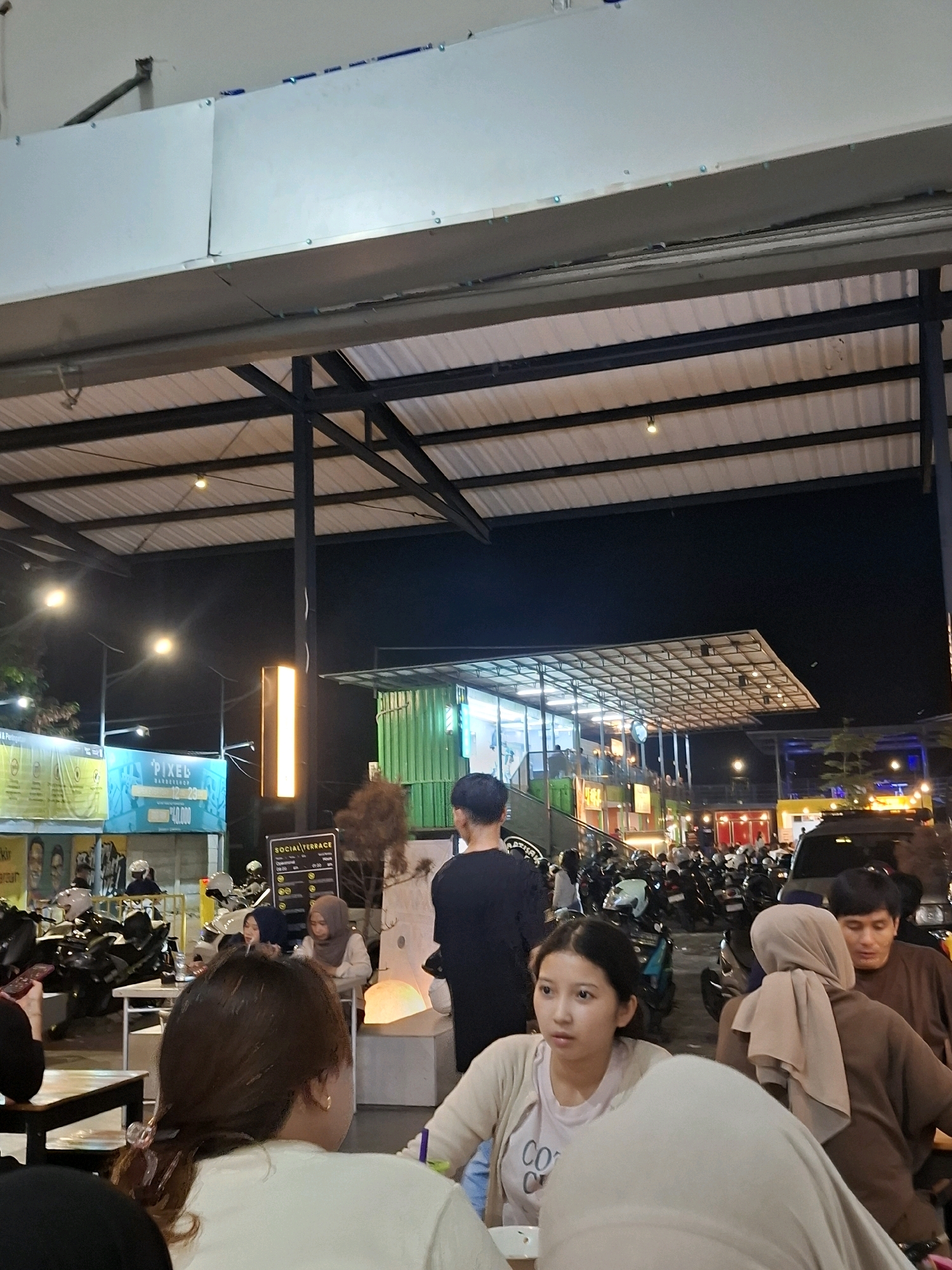 This is a medium shot, taken at night, of a busy outdoor food court in Cianjur, Indonesia. Several people are seated at tables, with many more standing or walking around. A variety of motorcycles are parked in the background, suggesting this is a popular gathering spot. The area is covered by a large, corrugated metal roof supported by metal beams, and illuminated by overhead lights and signage. In the foreground, two young women are seated at a table, facing the camera. One is wearing a light-colored cardigan over a t-shirt, and the other is wearing a light-colored shirt. To their right, a man and two women wearing hijabs are also seated at tables, interacting with each other. The overall atmosphere appears lively and social, with people enjoying food and company.