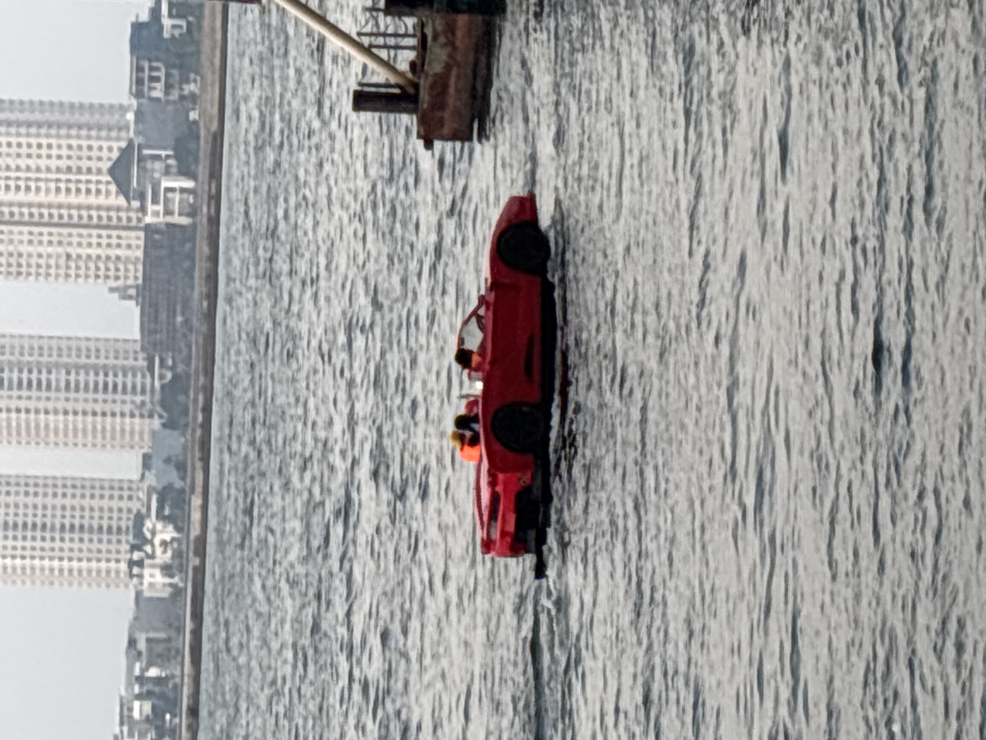 A red, amphibious sports car with two occupants wearing life vests is navigating through choppy water. The car has a low profile, and its wheels are submerged, suggesting it is operating in a watercraft mode. The two individuals are seated in the front seats, with one appearing to be steering. In the background, a city skyline is visible, characterized by numerous tall buildings and a pier structure to the right of the frame. The sky is overcast. The scene appears to be located in North Jakarta, Indonesia, given the architectural style of the buildings and the presence of a harbor or coastal area.