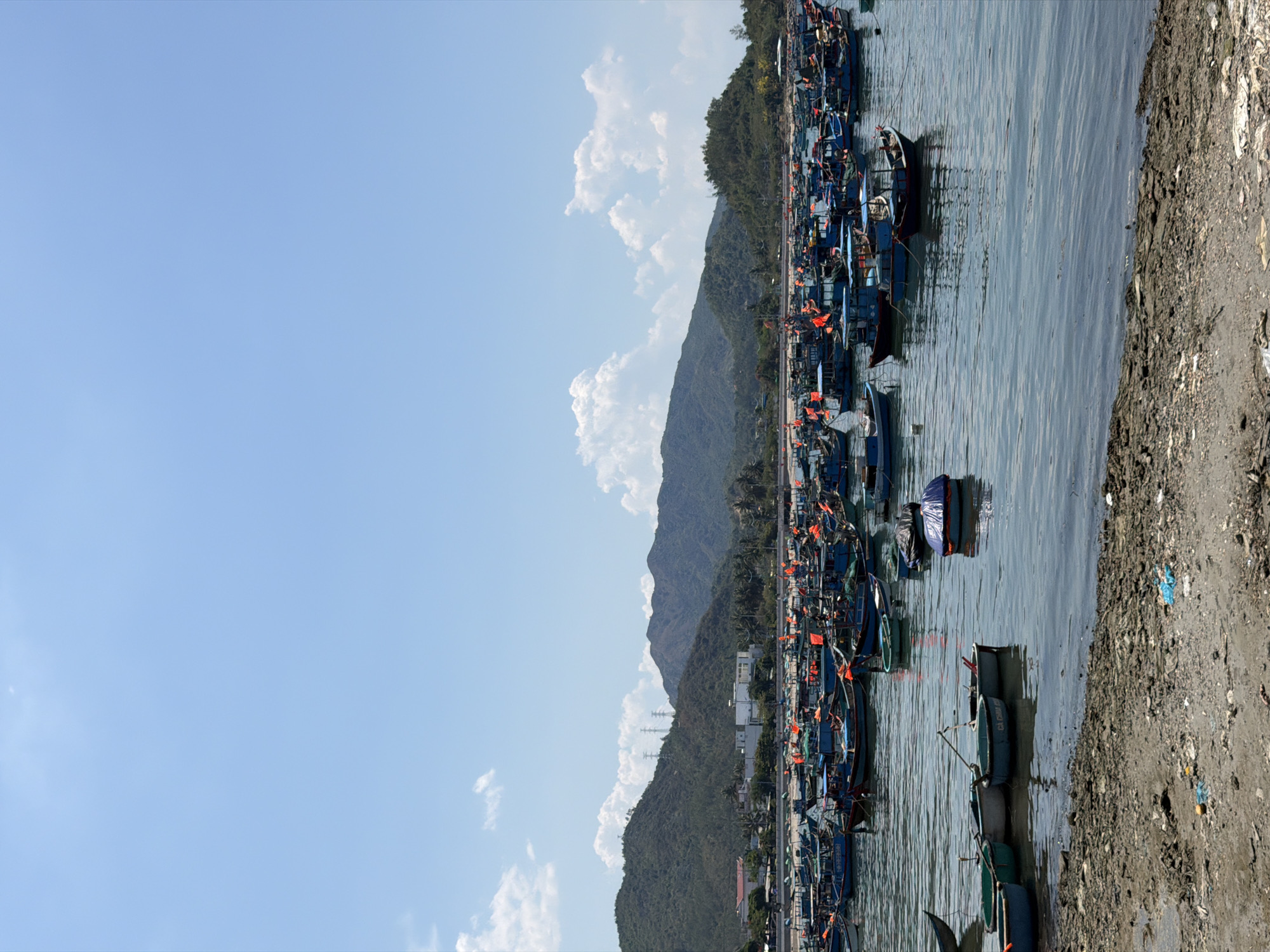 The image shows a harbor in Vĩnh Lương, Vietnam. A multitude of fishing boats are docked along the shore. Mountains rise in the background, partially obscured by clouds. The sky is blue. A small stretch of beach, covered in dark-colored dirt and debris, is visible in the foreground.