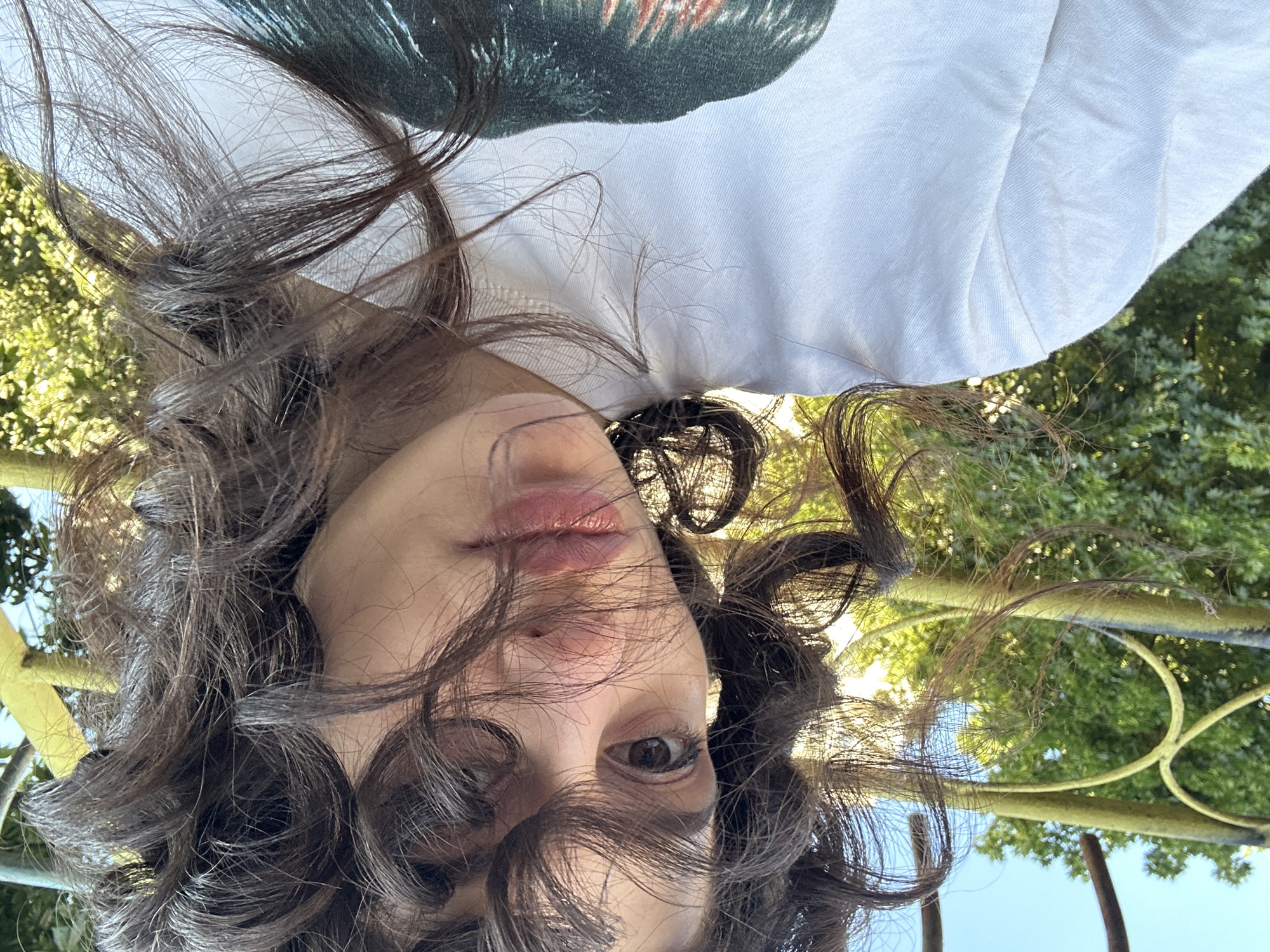 A close-up, outdoor, eye-level, selfie-style photograph shows a young person from the chest up. They have dark, curly hair that partially obscures their face. Their left eye is visible, with dark eyelashes and brown iris. Their lips are slightly parted and appear to have a pinkish tint. They are wearing a white t-shirt with a graphic print on the chest, the details of which are not clearly discernible. The background consists of lush green foliage and a portion of a yellow and brown metal structure, possibly a fence or railing, against a clear blue sky. The scene is illuminated by natural sunlight, casting shadows and highlighting the textures of the hair and clothing. The setting appears to be a park or garden.