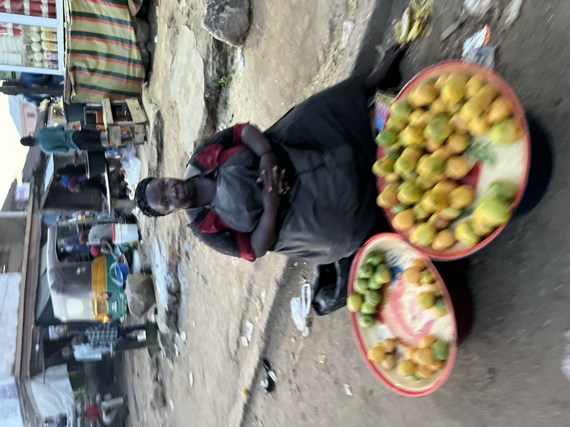 The image captures a vibrant street market scene, characteristic of an urban environment in Jos, Nigeria, during the daytime. The primary subject is a woman, likely a vendor, seated on the ground in the foreground. She is dressed in a dark garment, possibly a dress or skirt, over a red and black striped top, with her hands clasped in her lap. Her dark hair is neatly styled, and she looks towards the viewer with a calm, neutral expression.
Beside her are two large, round, shallow trays brimming with fresh fruit, predominantly mangoes, displaying a range of green and yellow hues. The larger tray holds a greater quantity, while the smaller one contains fewer fruits. A cluster of bananas is also visible near the fruit.
Behind the woman, the market unfolds with further activity. To her immediate left, a stall with a distinctive striped awning (green, red, and black) displays merchandise. Further into the background, other market structures, indistinct figures of people moving about, and what appears to be a "Keke Napep" (a motorized tricycle taxi commonly used for transport in Nigeria) can be seen. The ground is a mix of uneven dirt and concrete, with some scattered litter, typical of a busy, open-air market. The natural lighting suggests it is daytime, possibly morning or late afternoon. No discernible text or signage is visible within the frame. The scene conveys an authentic depiction of daily commerce and life in a bustling Nigerian marketplace.