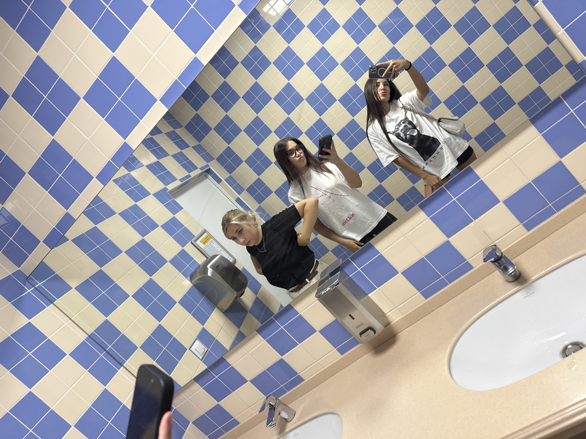 Three young women are taking a mirror selfie in a restroom with a blue and cream checkered tile pattern. The woman on the left is wearing a black top, the middle woman is wearing a white t-shirt with text on it and glasses, and the woman on the right is wearing a white t-shirt with a dog graphic and a small silver purse. One of the women is holding a phone to take the picture, and another is holding a phone and possibly a tablet. In the background, there are sinks, faucets, a soap dispenser, and a hand dryer mounted on the wall. The overall environment appears to be a public restroom. There is no indication of the location being in Bardar, Moldova.