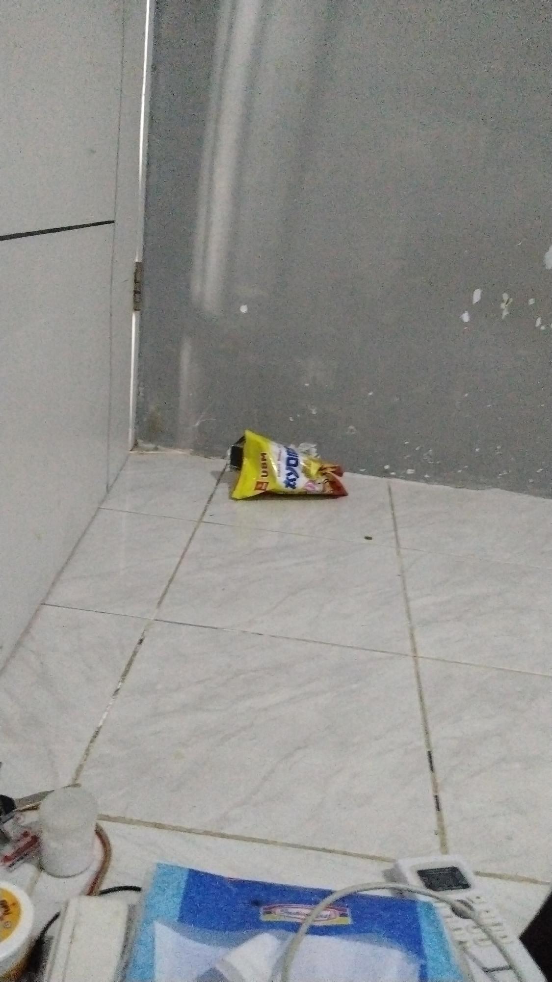 An unsealed, yellow, rectangular bag of XYDIX brand chewing gum lies on a tiled floor in a corner formed by two walls and a white door frame. The bag is partially open and appears to be the primary subject of the image. The floor is tiled with light-colored squares, and the walls are painted in a neutral, slightly textured gray. In the foreground, out of focus, are several objects including a white cylindrical object, a portion of a blue binder, and a white electronic device that resembles a cordless phone. The scene appears to be indoors, possibly in a residential or commercial space.