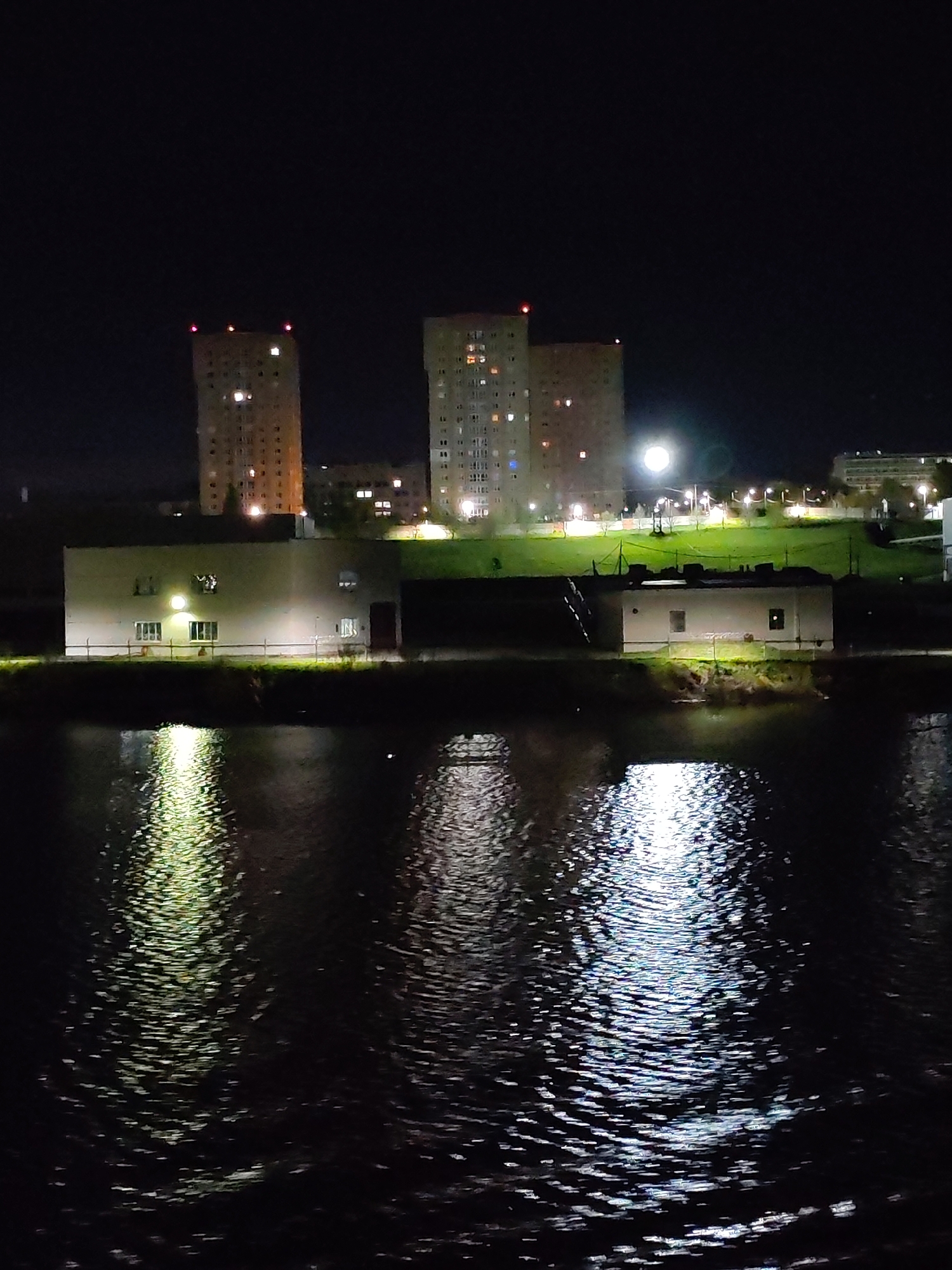 The image shows a nighttime view of a cityscape reflected in a body of water, likely a river or lake. In the background, there are three tall buildings, possibly residential towers, with lights visible in some of the windows and red lights on the roofs. A low, rectangular building is positioned in the middle ground, with lights emanating from its windows and reflected in the water. To the right, another small, rectangular building is present. The lighting suggests it is a night shot, with light sources being primarily from buildings, streetlights, and the moon or a bright light source in the background. The water surface is dark and rippled, creating distorted reflections of the buildings and light sources. The general location is in the city of Unknown, Unknown.