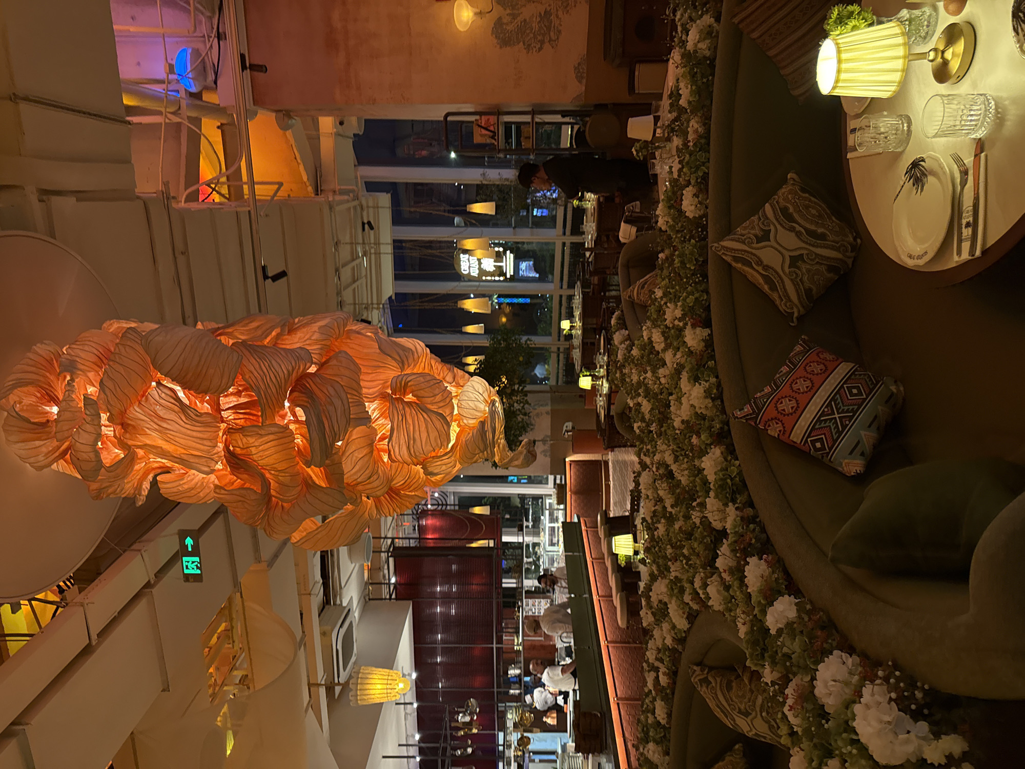 The image depicts the interior of a restaurant in Yiwu, China, with a distinctive opulent and botanical design. Dominating the center is a large, elaborate chandelier made of layered, translucent orange material, resembling petals or leaves, casting a warm glow. The restaurant features plush, olive-green curved seating areas adorned with numerous white flowers and greenery, creating an immersive garden-like atmosphere. Scattered across the seating are decorative pillows with patterned designs.
In the foreground, a round table is set for dining, complete with plates, cutlery, glassware, and a small yellow table lamp. In the mid-ground, a long counter separates the dining area from the kitchen. Several staff members, dressed in white chef attire, are visible working behind the counter, interacting with each other. The kitchen area is characterized by red-tiled walls and stainless steel equipment.
Further into the background, large windows offer a view of the city at night, with streetlights and building lights visible. A sign with the text "GREAT ATLANTIC" and a palm tree graphic is illuminated outside. The overall lighting in the restaurant is warm and ambient, with various lamps and spotlights highlighting the decor and creating an intimate setting. An illuminated green "exit" sign is visible on the ceiling in the upper left quadrant of the image. A person in dark clothing stands near the window in the background, looking out. The floor is not clearly visible due to the extensive floral arrangement.