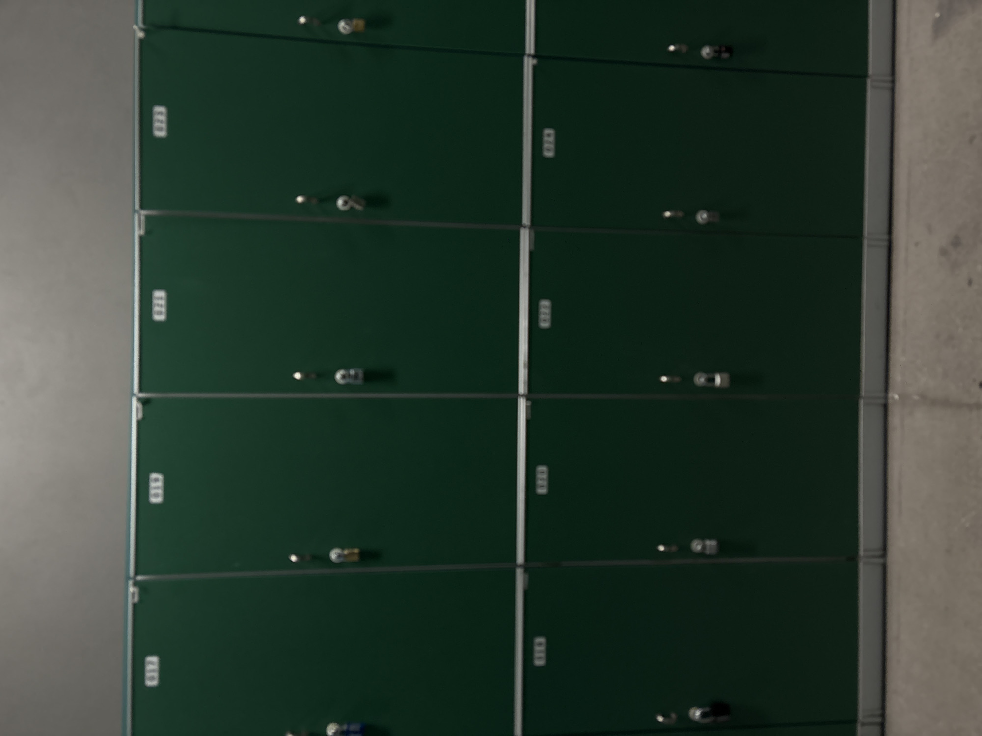 A bank of green metal lockers stands against a light gray wall and a concrete floor. The lockers are arranged in a grid pattern, with two columns and several rows visible. Each locker has a small number plate and a handle or locking mechanism. The location is unclear and cannot be determined from the image.