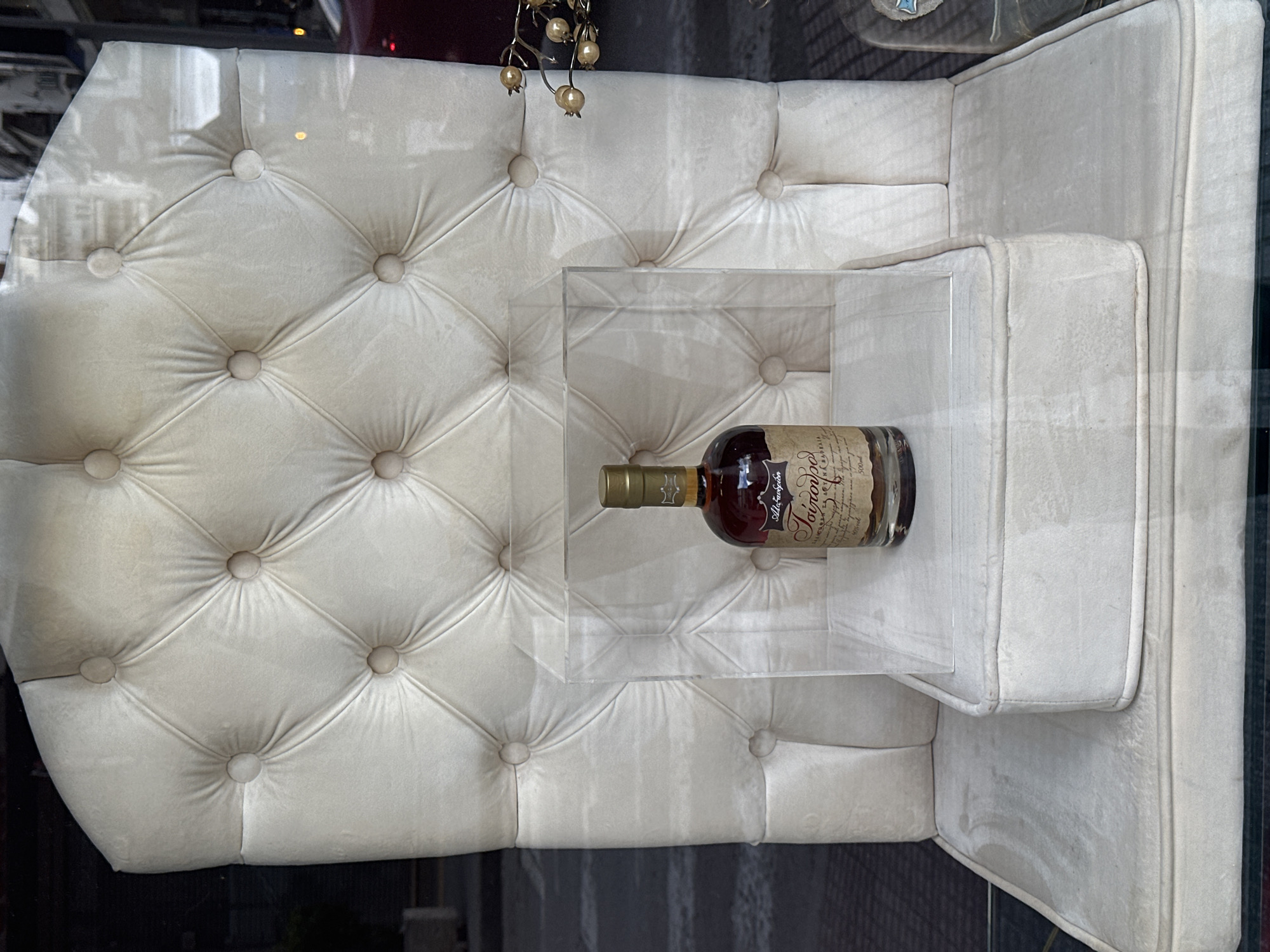 A bottle of "Tsipouro Alexandrion" is displayed on a clear acrylic stand placed on a tufted, off-white or beige chair or love seat. The background suggests the reflection of an urban scene, potentially a street. The location is Serres, Greece, inferred from the brand of Tsipouro being Alexandrion. The bottle is the primary object of focus, with the chair and display stand acting as supporting elements for the product display.