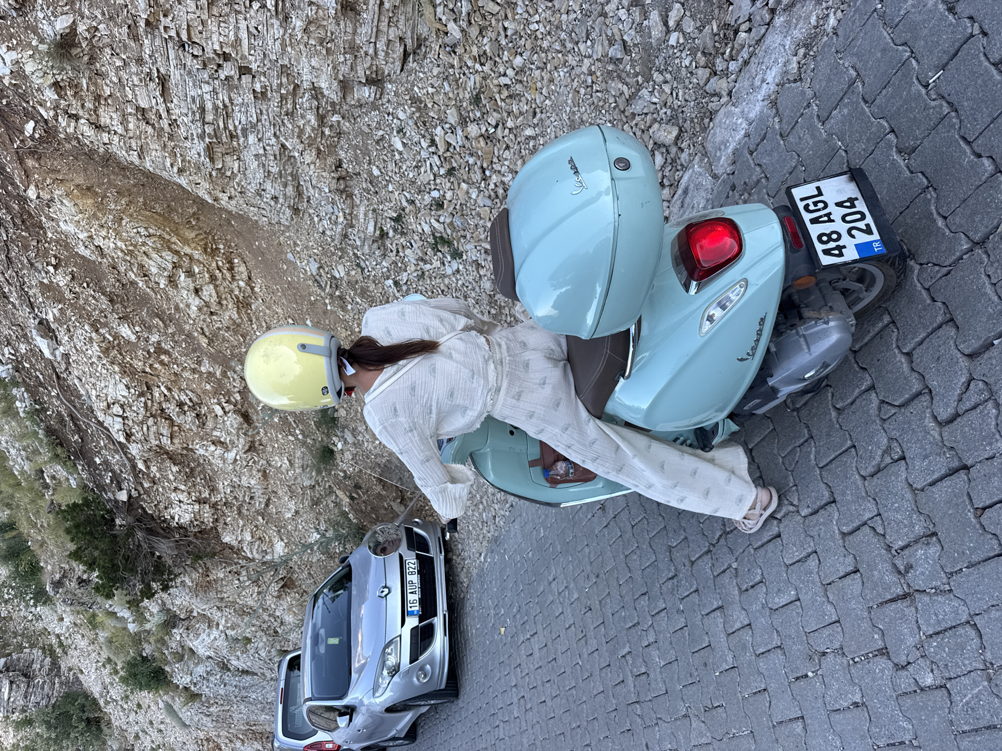 A person wearing a light-colored outfit and a yellow helmet sits on a light blue Vespa scooter, facing away from the camera. The scooter has a light blue storage box mounted on the back. To the left of the scooter, a silver car is parked. The scene appears to be on a paved road with cobblestone-like bricks, situated next to a rocky, mountainous embankment. The license plate on the scooter reads "48 AGL 204 TR." The license plate on the car reads "16 AUP 822." The environment suggests a location in Fethiye, Turkey, likely a coastal or mountainous area with significant rock formations.