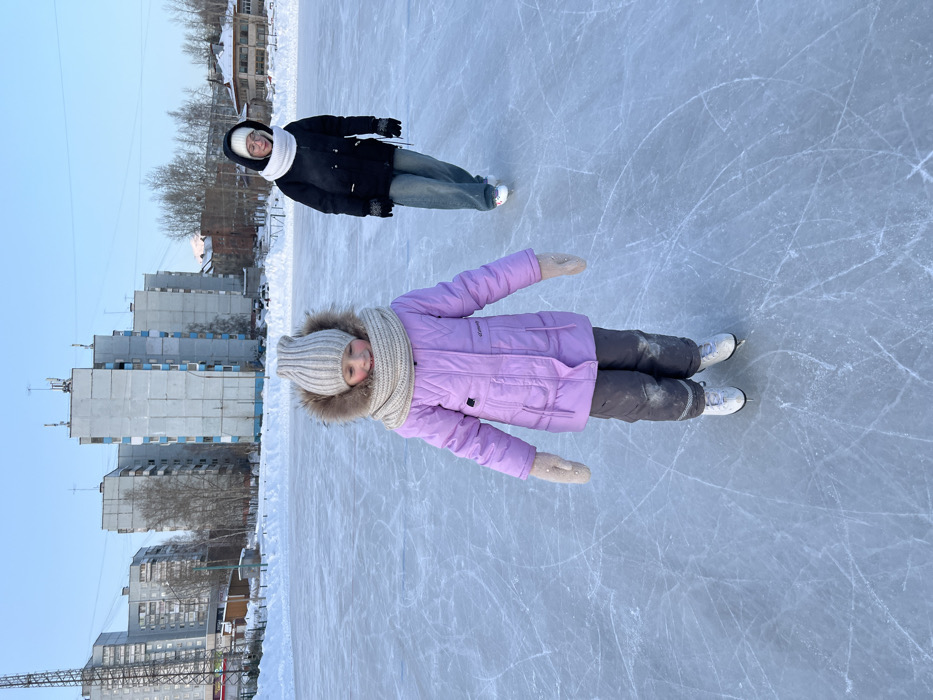 Two individuals are observed on an outdoor ice skating rink in Ust-Ilimsk, Russia. The foreground features a young child, estimated to be between 5-8 years old, standing center-left on white figure skates. This child wears a lavender winter coat, dark grey snow pants with a reflective band on the left knee, light beige mittens, a ribbed beige knit hat with a fur pom-pom, and a matching beige knit infinity scarf. The child's head is tilted slightly down and to the left, exhibiting a subtle smile. Traces of snow or dirt are present on the child's lower pants.
In the background, positioned to the right of the center, an older individual, likely a teenager, is seen wearing white ice skates. This person is dressed in a black hooded winter jacket, light blue wide-leg jeans, black gloves, a white knit infinity scarf, and clear-framed glasses beneath a white knit hat or hood. This individual appears to be gliding, facing slightly right with the head turned towards the left, displaying a faint smile.
The ice rink surface exhibits numerous skate marks and scratches. Surrounding the rink is a substantial layer of snow. In the background, several multi-story residential buildings, consistent with Soviet-era architecture featuring grey/light blue concrete and light blue balcony accents, are visible under a clear, light blue sky. Bare deciduous trees and further snow-covered ground are also present between the rink and the buildings. The scene is illuminated by natural daylight, indicating a clear winter day.