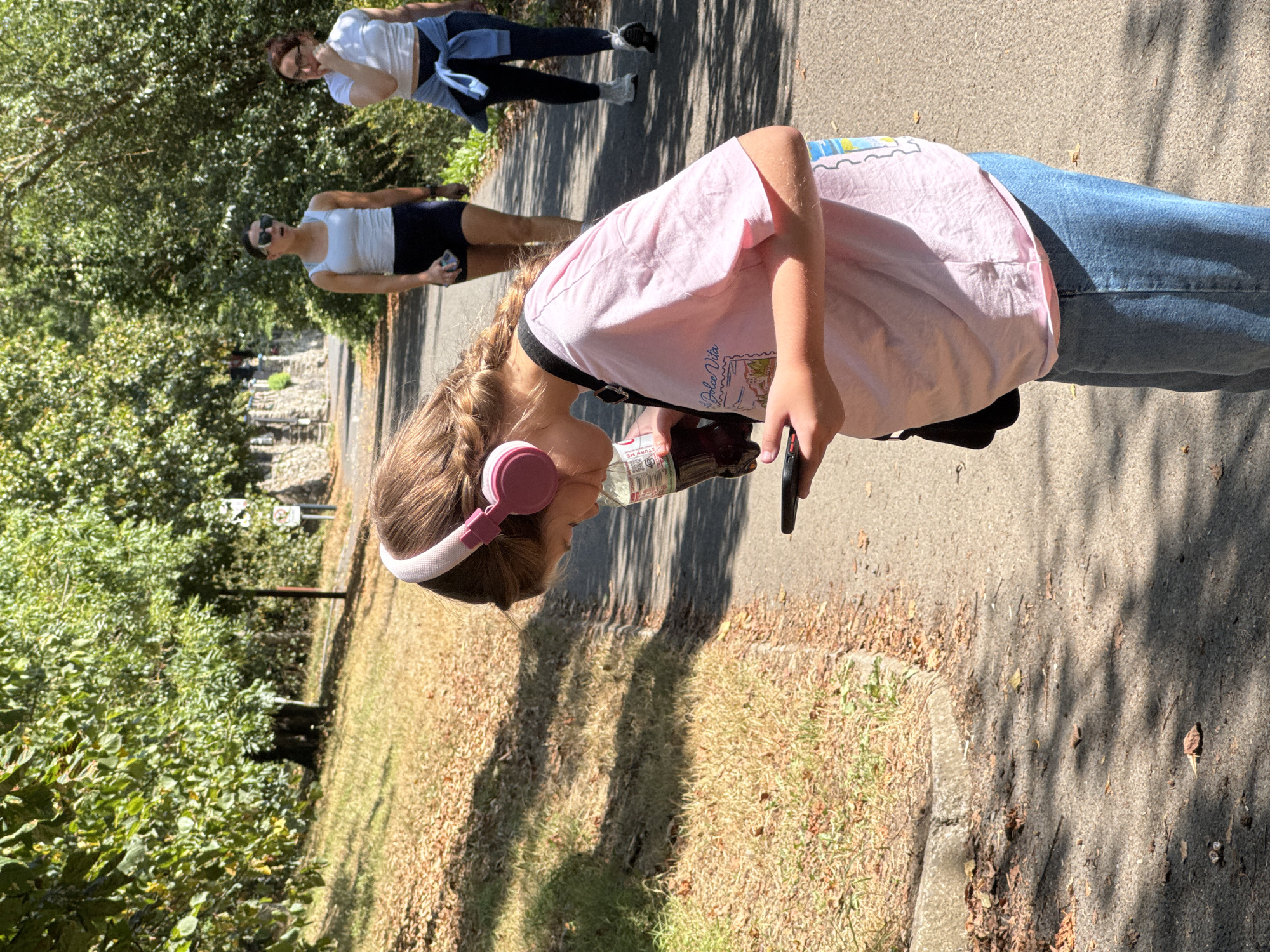 The image shows three people walking on a paved path outdoors. The foremost subject is a young person wearing a pink t-shirt, blue jeans, and pink headphones. They are holding a smartphone and a bottle of water. Behind them, two adults are walking. The person on the left wears a white tank top and dark shorts, with sunglasses on. The person further back is wearing a white t-shirt and blue leggings, with a blue jacket tied around their waist and glasses. The scene is a park or wooded area with trees and dry grass. Shadows from the trees are cast on the path, suggesting it is daytime, likely late morning or early afternoon given the angle of the sun. The overall mood appears casual and relaxed. Visible text on the water bottle includes "VODKA" and other smaller print, suggesting it might be a novelty item rather than an actual alcoholic beverage. The location is identified as Clonmel, Ireland, which might imply a temperate climate.