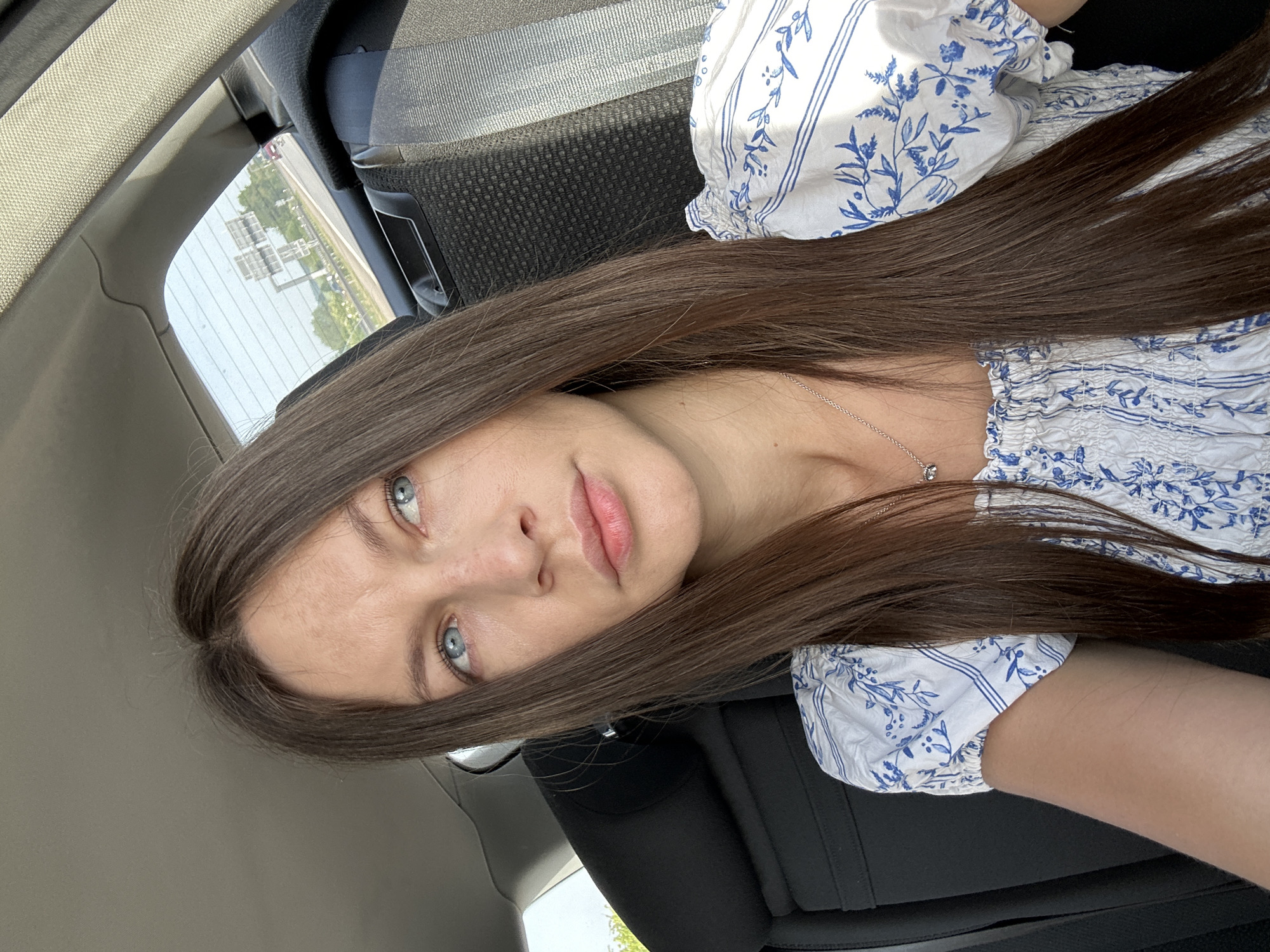The image shows a woman with long brown hair and blue eyes in a car. She is wearing a white and blue patterned top and a silver necklace. Outside the car window, a road sign is partially visible. The location is Rechtenbach, Germany.