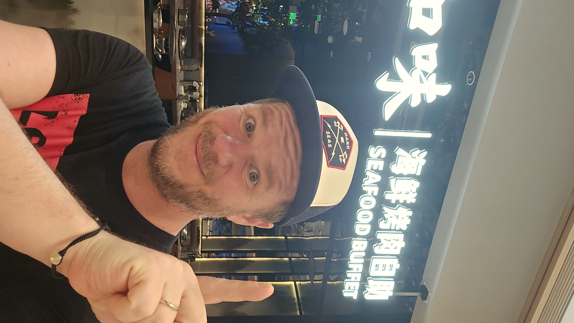 A man with light skin, a beard, and blue eyes is in the foreground, wearing a black t-shirt, a baseball cap with a patch that says "DARK SEAS", and a bracelet. He is looking at the camera and pointing his right index finger upwards. In the background, a neon sign reads "SEAFOOD BUFFET" in English, and Chinese characters. The sign is lit up in white, and the background is dark. The man is likely inside a restaurant in the city of Xiaoshan District, China.