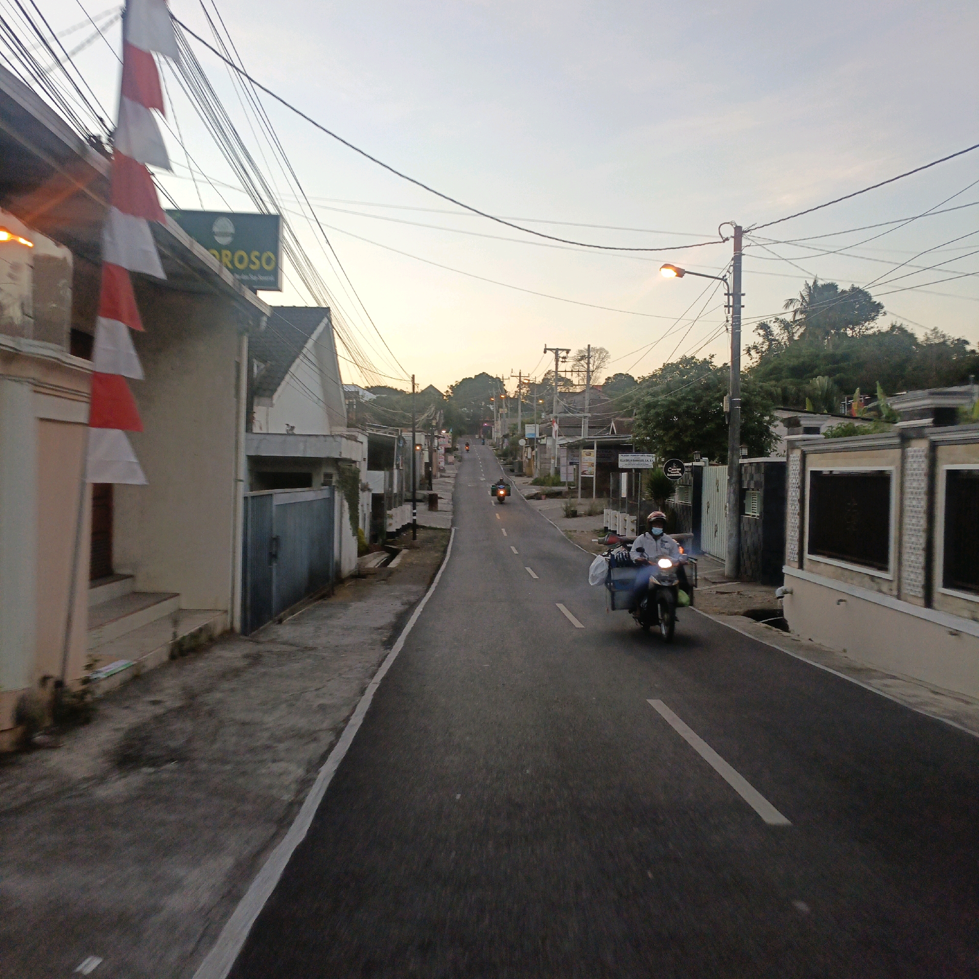 The image depicts a street scene in Salatiga, Indonesia, during what appears to be dusk or early dawn, with a softly lit sky. A paved road with white lane markings runs through the frame, bordered by buildings and vegetation. On the left side, a building with a cream-colored facade and a dark-colored gate is visible, adorned with red and white Indonesian flags. Further down the street, several buildings with varied architectural styles line the road.
A prominent feature is the network of utility wires and poles crisscrossing the sky. A street lamp is illuminated on the right side of the road. Two motorcycles are the primary subjects in motion. One motorcycle with a sidecar, driven by a person wearing a light-colored shirt and a blue face mask, is moving towards the viewer on the right side of the road. Another motorcycle, with its headlight on, is moving away from the viewer further up the street. The general atmosphere suggests a quiet residential or mixed-use area in the city.