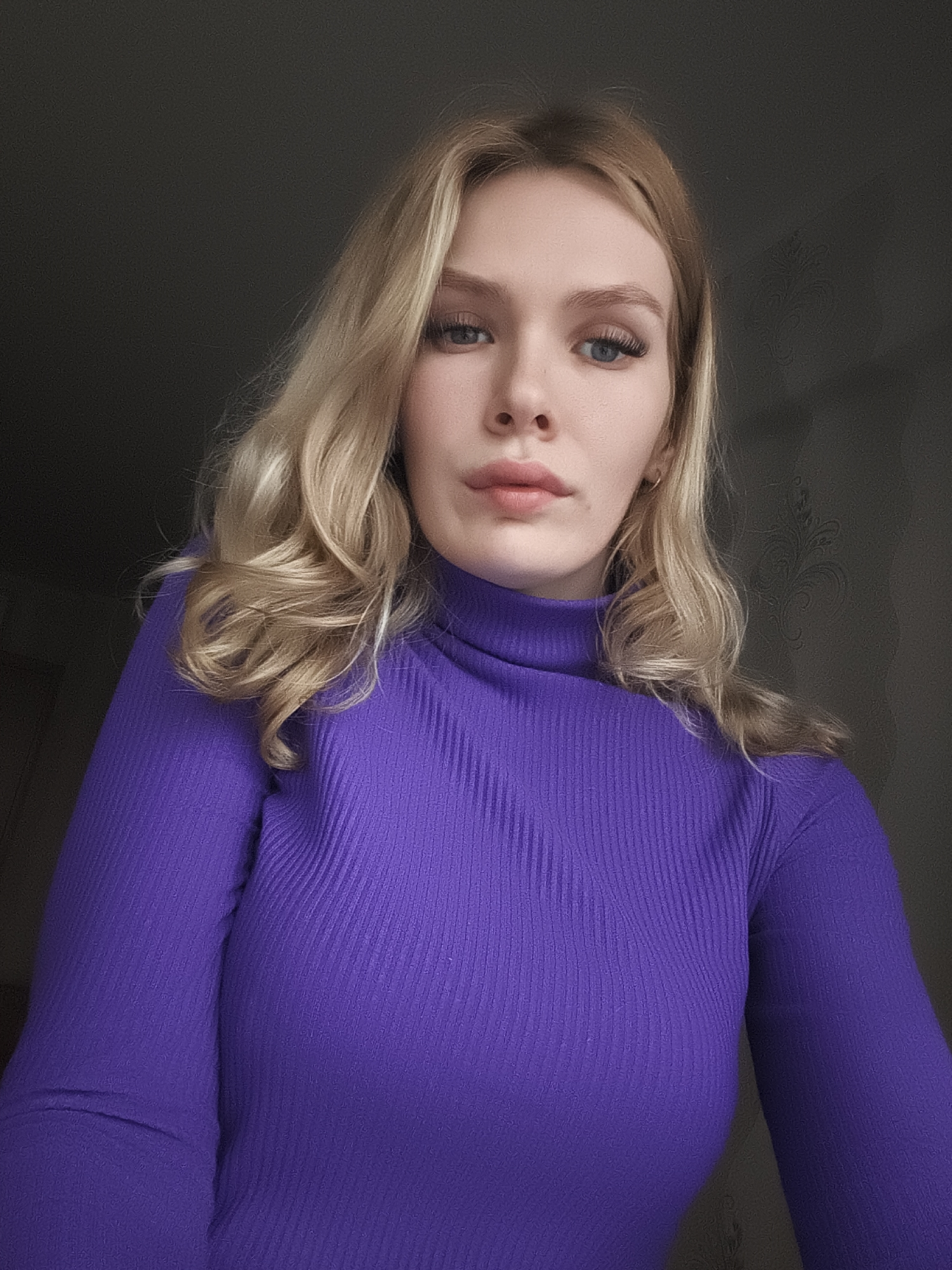 A portrait image features a young woman with long, wavy blonde hair and blue eyes, looking directly into the camera. She wears a form-fitting, ribbed purple turtleneck long-sleeved top. Her expression is neutral and direct, with a subtle hint of pink lipstick and enhanced eyelashes visible. The lighting comes from the front, highlighting her features.
The background is dark and indistinct, consisting of a plain wall on the left and a darker wall with a subtle, dark patterned wallpaper on the right. She appears to be posing for the photograph, likely taken indoors. No specific time of day or weather can be determined from the shot. There are no discernible location cues within the image itself, apart from the general indoor setting. No text is visible. The location context for this image is Izhevsk, Russia.