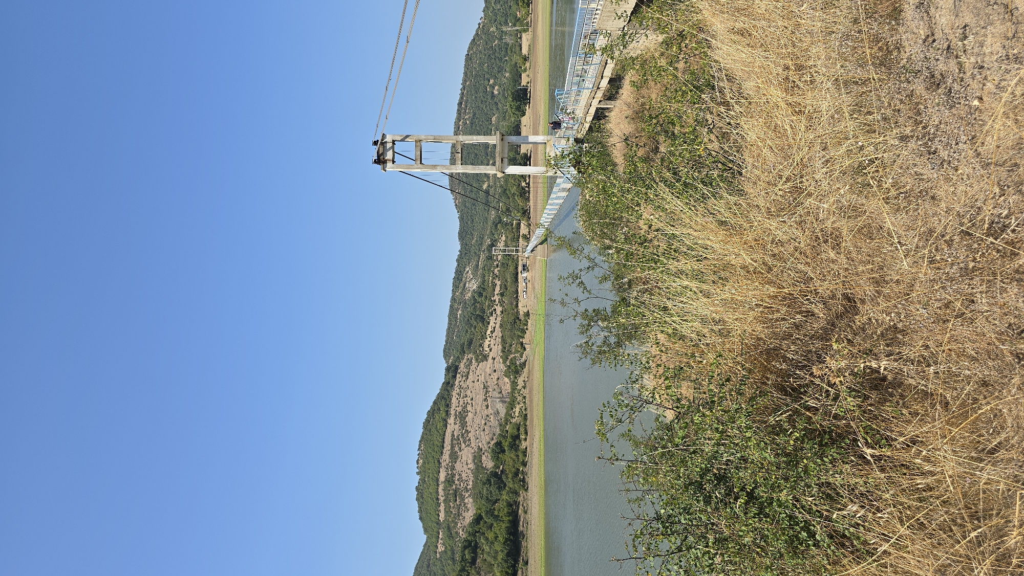 In the city of Lisitsite, Bulgaria, a suspension bridge spans a wide body of water. The bridge, with its concrete towers and steel cables, is illuminated by the bright, clear blue sky. The water reflects the blue sky, creating a serene atmosphere. On the bridge, a few people are visible, strolling and enjoying the view. The landscape surrounding the water is characterized by rolling hills covered in dry, golden grass and scattered green bushes, suggesting a warm, dry climate. The foreground of the image is dominated by dry, tall grass, adding a rustic touch to the scene.