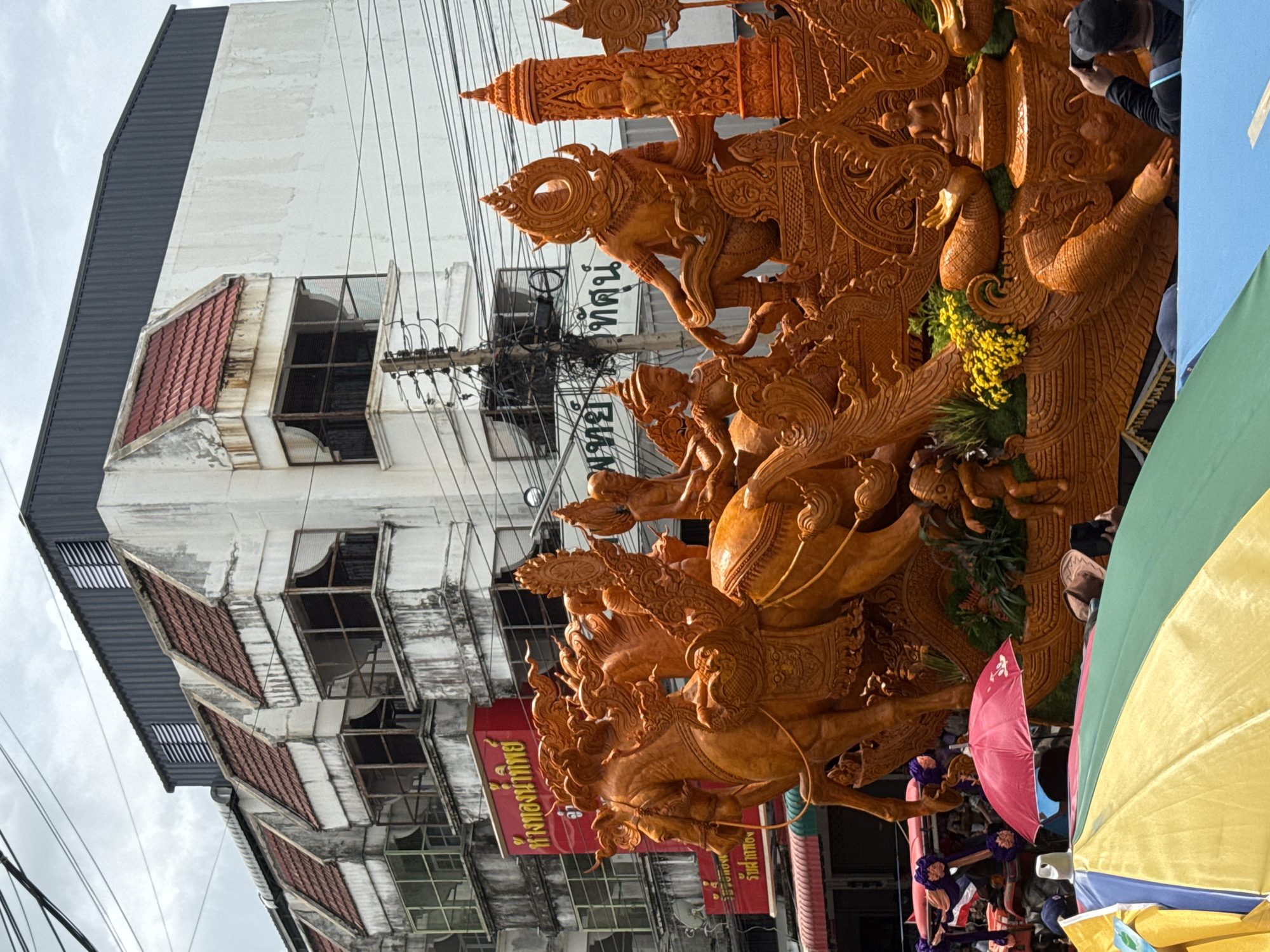 The image depicts a street scene in Prakhon Chai District, Thailand, during what appears to be a parade or festival. A large, intricately carved orange float, likely made of wax, dominates the foreground. The float features mythical creatures, including a multi-headed horse with riders and elaborate decorative elements. Behind the float, a row of multi-story buildings with weathered facades and red tiled roofs line the street. Overhead, power lines crisscross the sky, indicating an urban setting. In the lower portion of the image, the tops of colorful umbrellas and crowds of people are visible, suggesting a public gathering. One person in the lower right corner is taking a photograph of the float with a mobile phone.