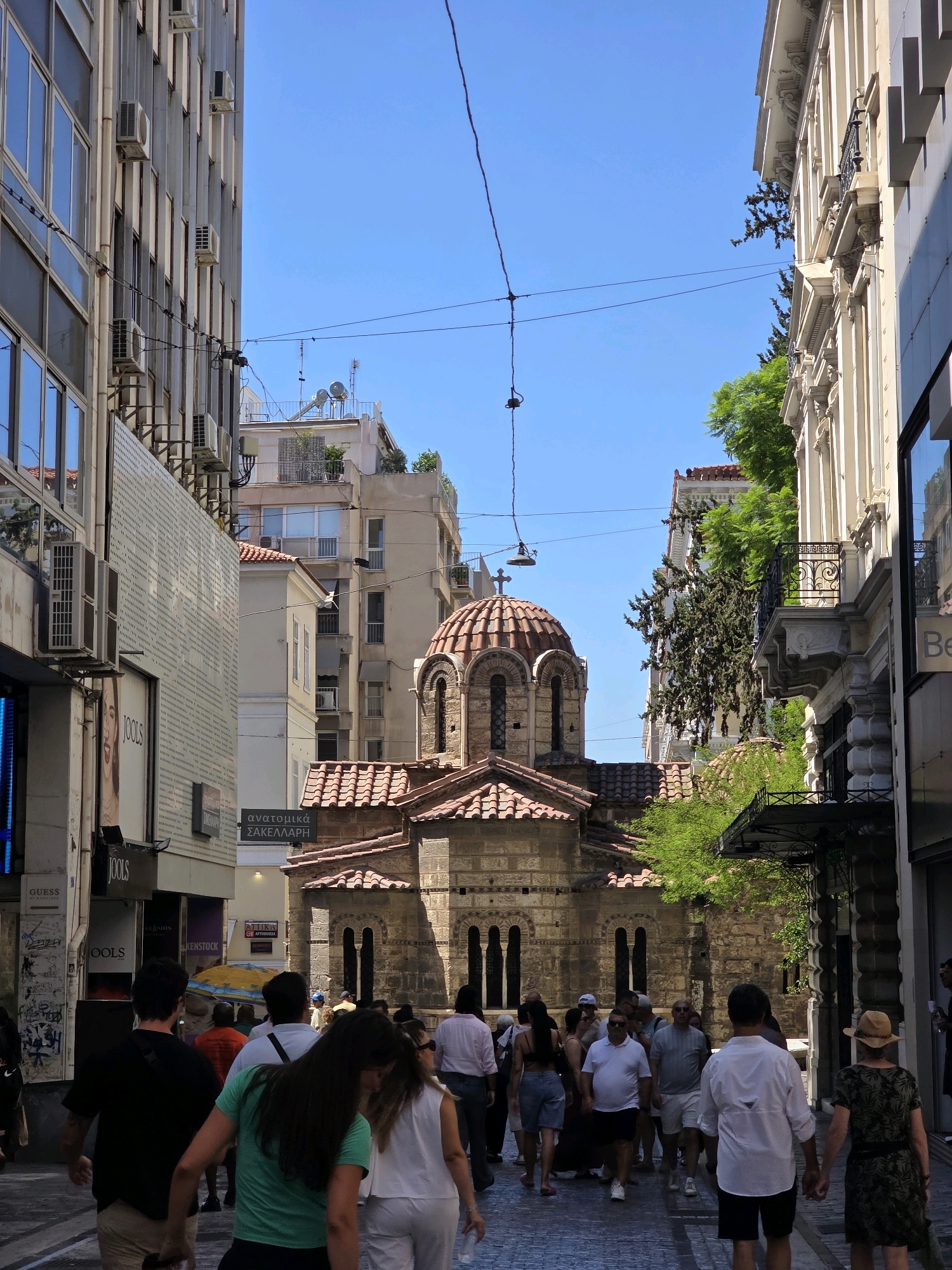 This is a street scene in Athens, Greece, featuring a small Byzantine church situated between tall, modern buildings. The church has a red-tiled roof and a dome, with stone walls and arched windows. A street runs through the frame, with several people walking along it. The buildings on either side of the street are several stories high, with one on the left appearing to be a commercial building with shops at street level, and the one on the right also displaying commercial signage. The sky is clear blue. The overall impression is a juxtaposition of historical architecture with the bustling city life of Athens.