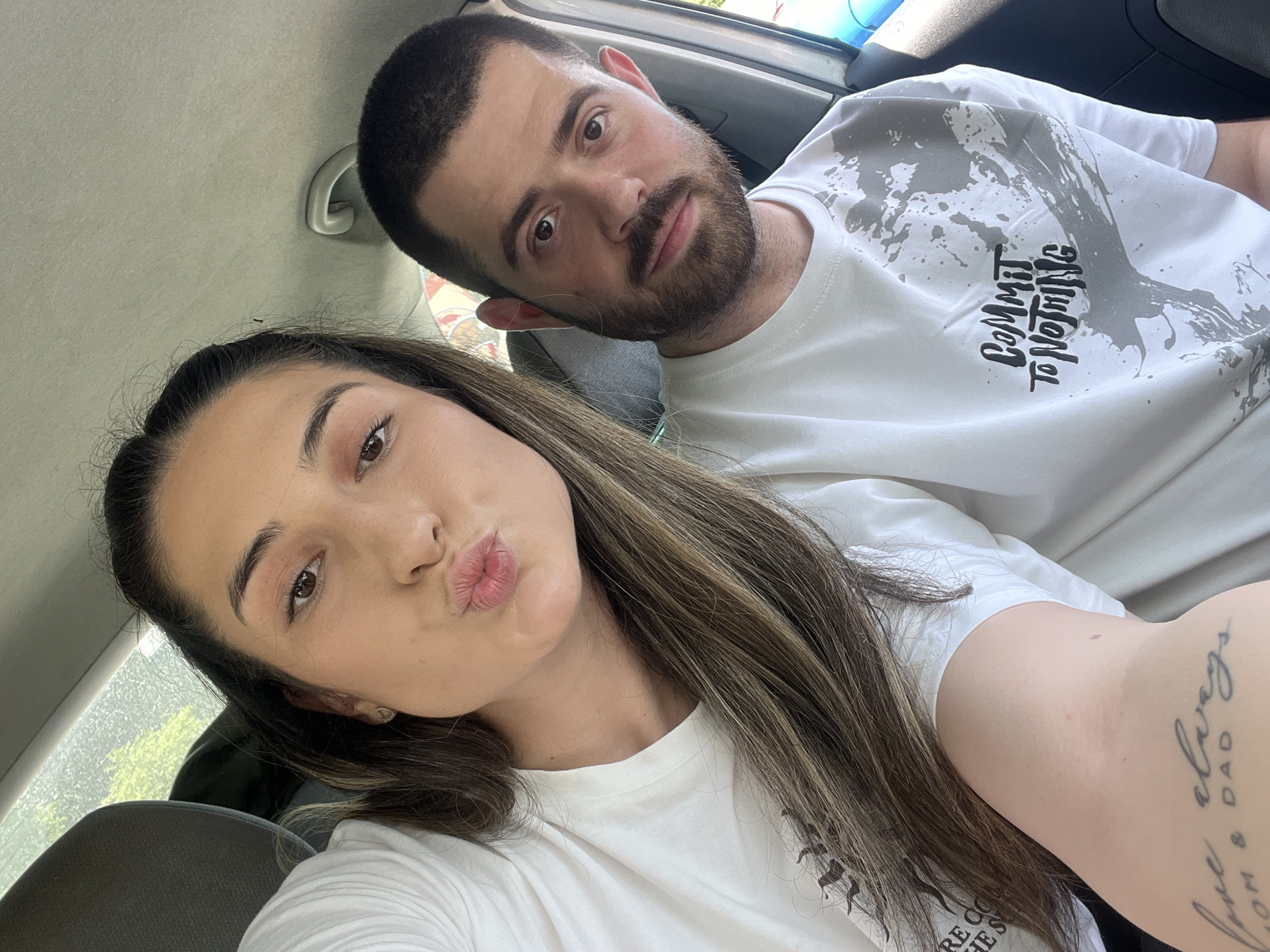 This image captures a selfie-style portrait of two individuals, a female and a male, inside a vehicle, presumably in Plovdiv, Bulgaria. The female, positioned prominently in the foreground on the left, has long brown hair with lighter streaks, partially pulled back. She is making a "kissing" face towards the camera, with her lips puckered. Her visible right eye is dark brown. She wears a plain white t-shirt, and her left forearm, visible in the lower left, displays a tattoo that reads "love always MOM & DAD" in script. A small, circular, gold-colored stud earring is visible in her right earlobe.
Behind and slightly to the right of the female, the male is seated, looking directly at the camera with a neutral expression. He has short dark hair and a dark beard. He is wearing a white t-shirt adorned with a distinctive grey splatter graphic and bold black text that reads "COMMIT TO NOTHING".
The interior of the vehicle is visible, including a grey headrest behind the female on the left, a light-colored (beige/grey) car ceiling/headliner, and a curved grab handle mounted on the ceiling above the male's head. Through the car's window in the upper background, blurred green foliage is visible, indicating an outdoor, daytime setting.