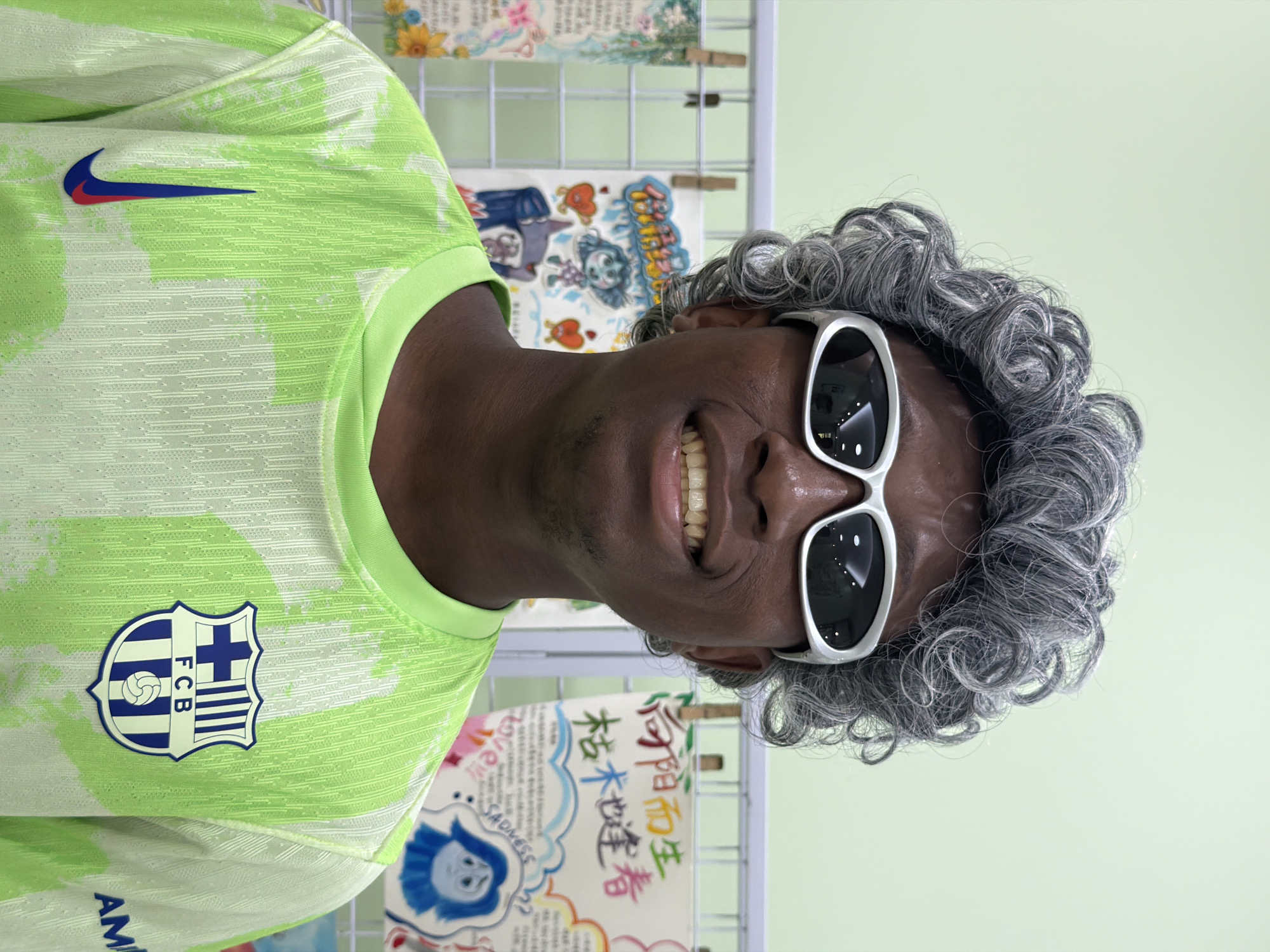 A dark-skinned person is captured in a close-up, waist-up portrait, smiling broadly with their head tilted slightly. They are wearing a vibrant lime green short-sleeved t-shirt featuring a subtle patterned texture and lighter green, abstract patches. On the wearer's right chest, a Nike swoosh logo in red and blue is prominently displayed, while on the left chest, an FC Barcelona crest with blue and red stripes, a cross, and a football is visible. Partial text "AM" can be seen near the bottom of the t-shirt.
The person is also wearing a voluminous, curly grey wig and stylish white sunglasses with dark lenses, which contribute to a playful and cheerful expression. Their broad smile reveals their teeth.
The background is an indoor setting with a light green wall. A white wire grid display stand holds several colorful drawings or posters, which appear to be children's art or educational materials. Visible text on these background items includes Chinese characters, such as "枯木逢春" (meaning "dry wood meets spring" or "rejuvenation") and "你阳光" (likely "you are sunshine"). English words like "SADNESS" and "LOVE" are also discernible on some of the posters. The overall atmosphere conveyed by the person's expression and attire, combined with the background, suggests a fun or themed event in a casual, possibly educational, environment in Jiangjin, China.