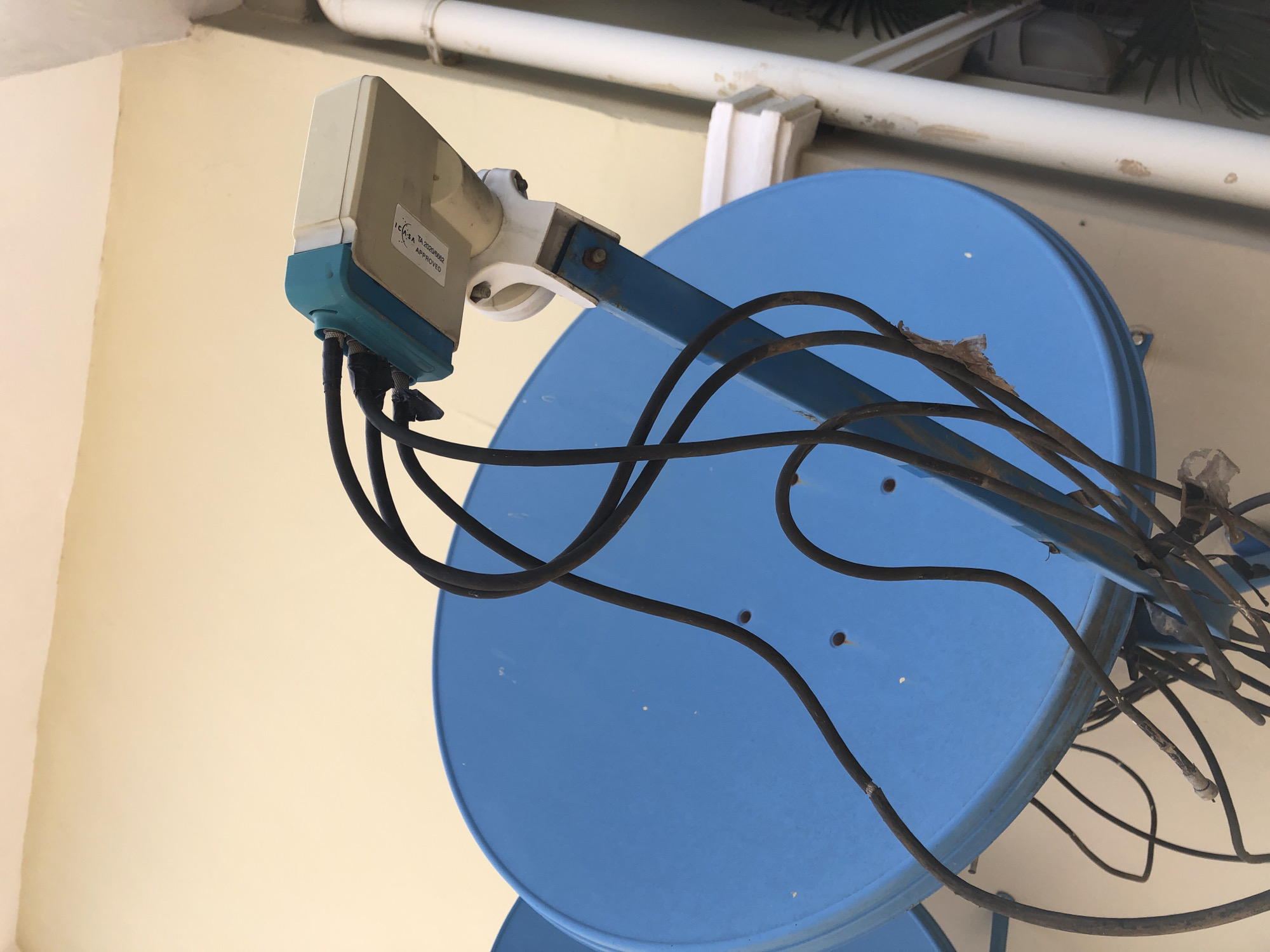 Two blue satellite dishes are mounted on a wall in Bar Jirgi Summa, Nigeria. The dish in the foreground is larger and appears to be the primary focus. It is supported by a blue metal arm that is attached to the wall. A white and teal colored LNB (low-noise block downconverter) is affixed to the arm, connected to multiple black coaxial cables. Some of these cables appear to be frayed and taped. The wall behind the dishes is painted a pale yellow. A white pipe runs vertically along the right side of the image. Some greenery is visible behind the pipe. The overall scene depicts typical outdoor satellite dish installation.