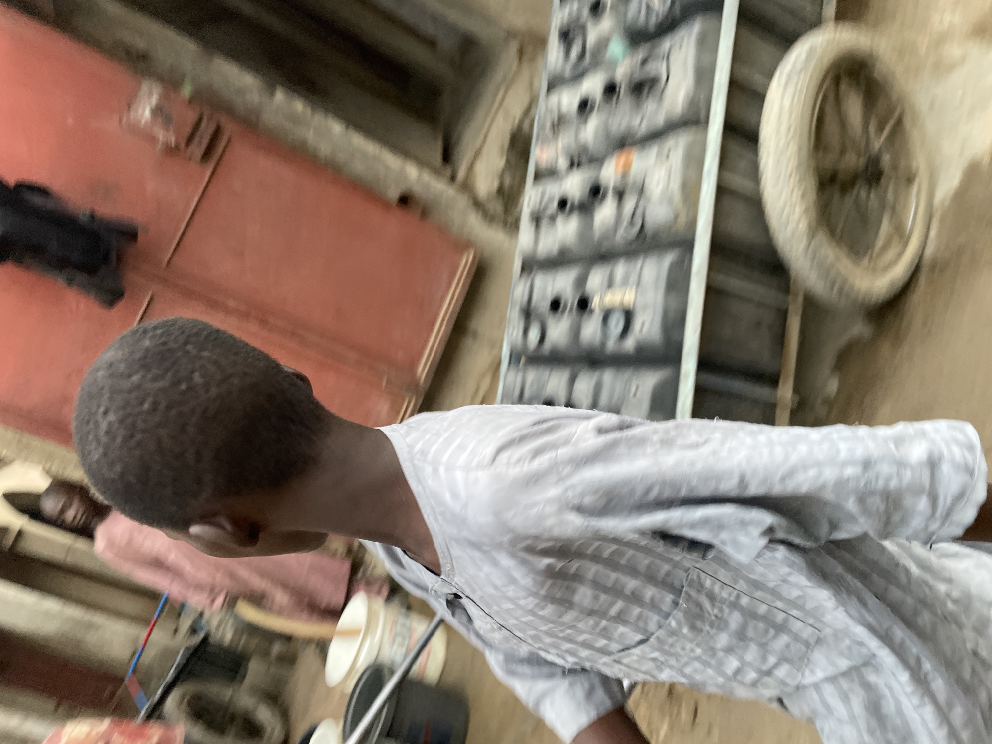 The image depicts a person with short, dark hair and wearing a light gray, striped shirt. In the background, there's a red door, a set of gray stacked objects with circular holes, and a tire mounted on a wheel. Additional indistinct figures and objects are partially visible, suggesting an indoor environment in a city.