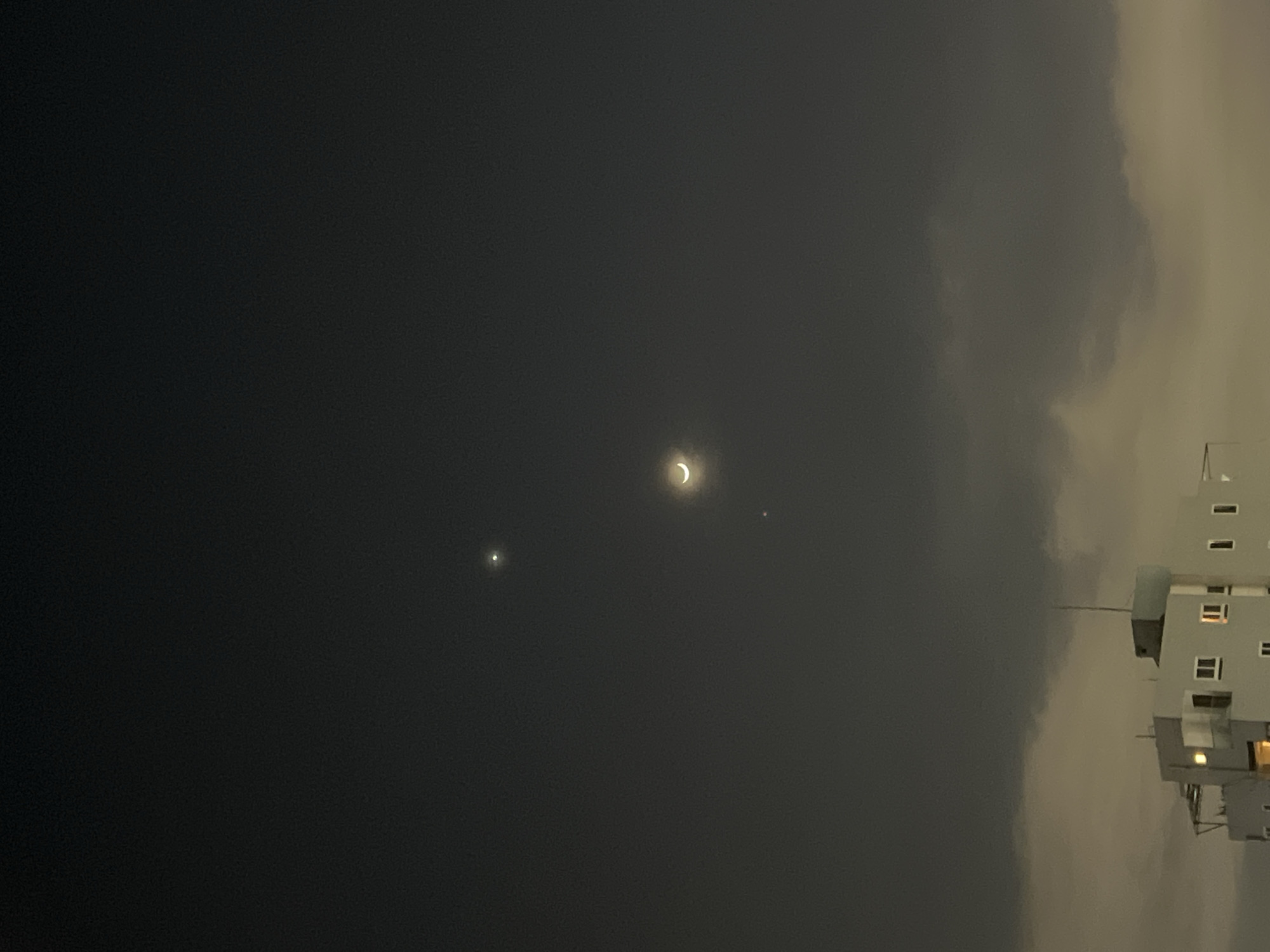 This image captures a nocturnal scene above a city, likely Đà Nẵng, Vietnam, given the context. The dark sky is punctuated by celestial bodies: a crescent moon, a bright point of light to its upper left, and another fainter point of light below the moon. These celestial objects are positioned against a hazy, dark sky.
Below the sky, a portion of a multi-story building is visible. It appears to be made of concrete and has several windows, some of which are illuminated. The building has a flat roof with some structural elements and a small antenna. The foreground is dominated by softly lit clouds, which are dense and obscure the lower parts of the scene, suggesting a slightly overcast or hazy atmosphere. There are no people or animals depicted in the image. The overall impression is one of a quiet, urban night with a clear view of the moon and planets.