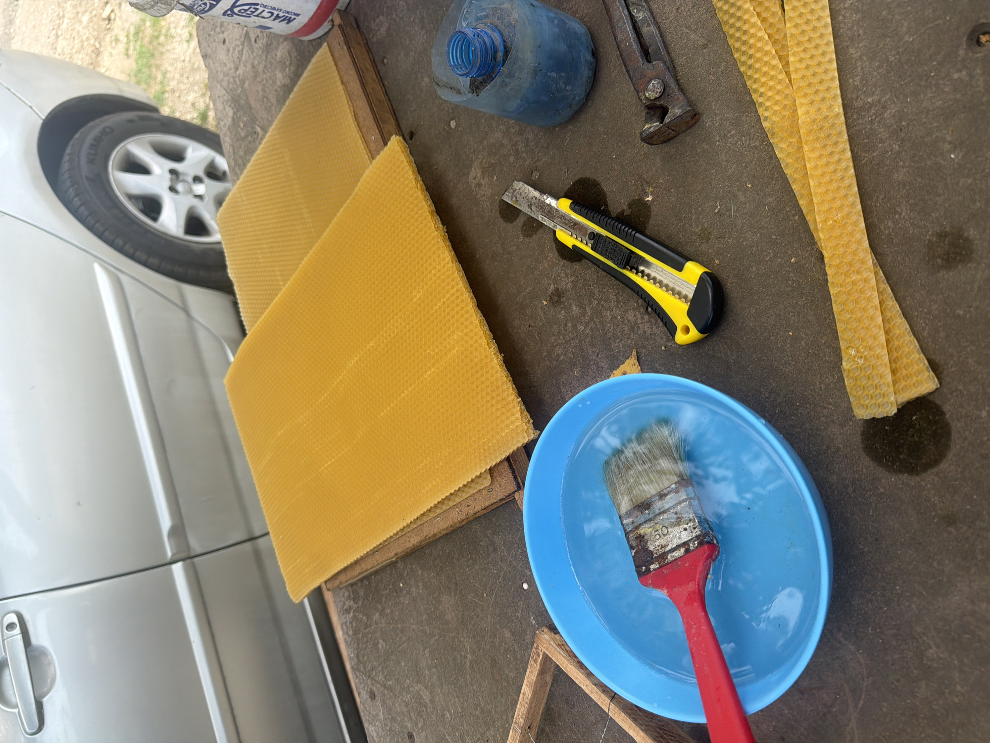 This image captures a close-up, overhead view of various items laid out on a textured, dark surface, likely a workbench or outdoor table. The scene suggests a do-it-yourself project involving beekeeping supplies, specifically honeycomb foundation sheets.
Dominating the left side of the frame is a portion of a silver vehicle, with its tire and part of the car door visible, indicating an outdoor setting. On the dark surface, two large, rectangular sheets of yellow honeycomb foundation are stacked on top of each other. To the right of the foundation, a bright yellow utility knife with a black handle lies open, ready for use. Further to the right, three narrow, ribbon-like strips of honeycomb foundation are arranged diagonally.
In the lower right quadrant, a bright blue plastic bowl contains water, with a paintbrush partially submerged. The paintbrush has a red handle and bristles that appear slightly worn. A small, wooden frame, possibly for beekeeping, is visible at the very bottom edge of the image.
In the upper portion of the image, a blue plastic bottle with a textured blue cap is positioned near a pair of old, rusty pliers. There is also a partially visible object with red and white cylindrical packaging, suggesting it might be some kind of glue or sealant. The overall impression is one of preparation or active engagement in a hands-on task. The lighting suggests it's daytime, and the presence of water in the bowl could indicate a need to dampen materials or clean tools. The setting appears to be outdoors, potentially in a rural area given the mention of Dolno Ablanovo, Bulgaria, although no specific landscape features are clearly visible. No people are present in the frame.
Visible text includes "KUMHO" on the tire and "MACTEP" on the cylindrical packaging in the upper left.