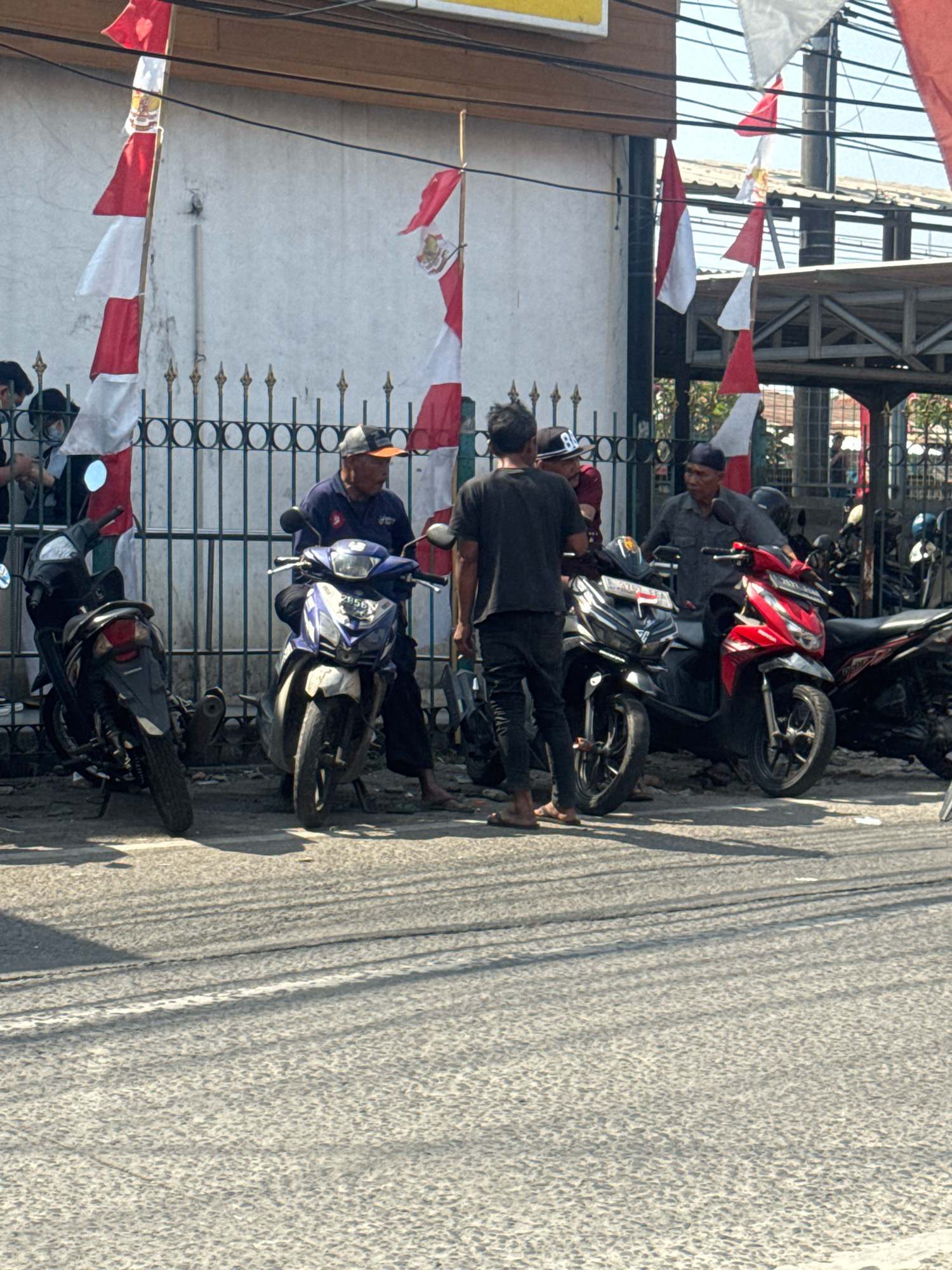 In Sukaraja, Indonesia, a group of men are gathered in front of a building with a white wall, possibly a shop or a house, on a street. Red and white Indonesian flags are prominently displayed, hanging from poles and draped along a black metal fence. Several motorcycles are parked on the asphalt road, some with their kickstands down.
In the foreground, a black motorcycle is parked on the left, and a blue and white motorcycle is parked next to it on the right. A man in a blue shirt and dark pants is sitting on the blue and white motorcycle, looking towards the right. Another man, wearing a black t-shirt and dark pants, is standing in front of a red motorcycle, facing the man in blue. Behind them, another man in a gray shirt and dark pants is seated on the red motorcycle. More motorcycles are visible further down the street, partially obscured by the parked vehicles. The overall scene suggests a casual gathering or waiting area. The lighting indicates it is daytime, with shadows cast on the road.