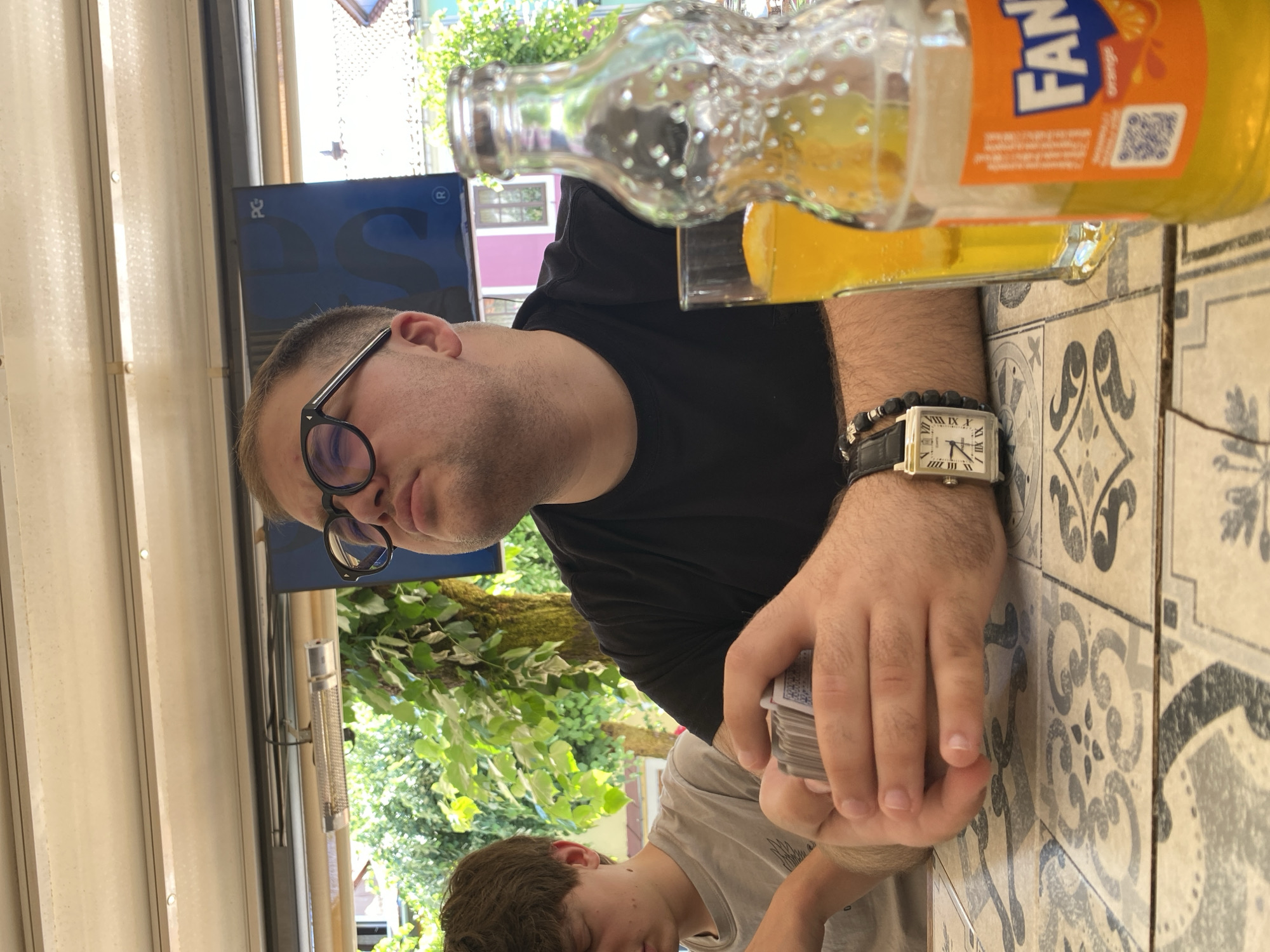 The image depicts two individuals seated at a table outdoors, possibly at a cafe or restaurant. The person in the foreground, a man with dark hair and glasses, is looking upwards and holding a deck of playing cards. He is wearing a black t-shirt and a watch. To his left, a younger person with lighter hair is seated and looking down. On the table are a glass of orange liquid and a bottle of Fanta. The background shows greenery and buildings, suggesting an urban outdoor setting. The table surface has a tiled pattern. The presence of a Fanta bottle and the general ambiance hints at a relaxed, casual setting, potentially in Cetinje, Montenegro, given the context provided.