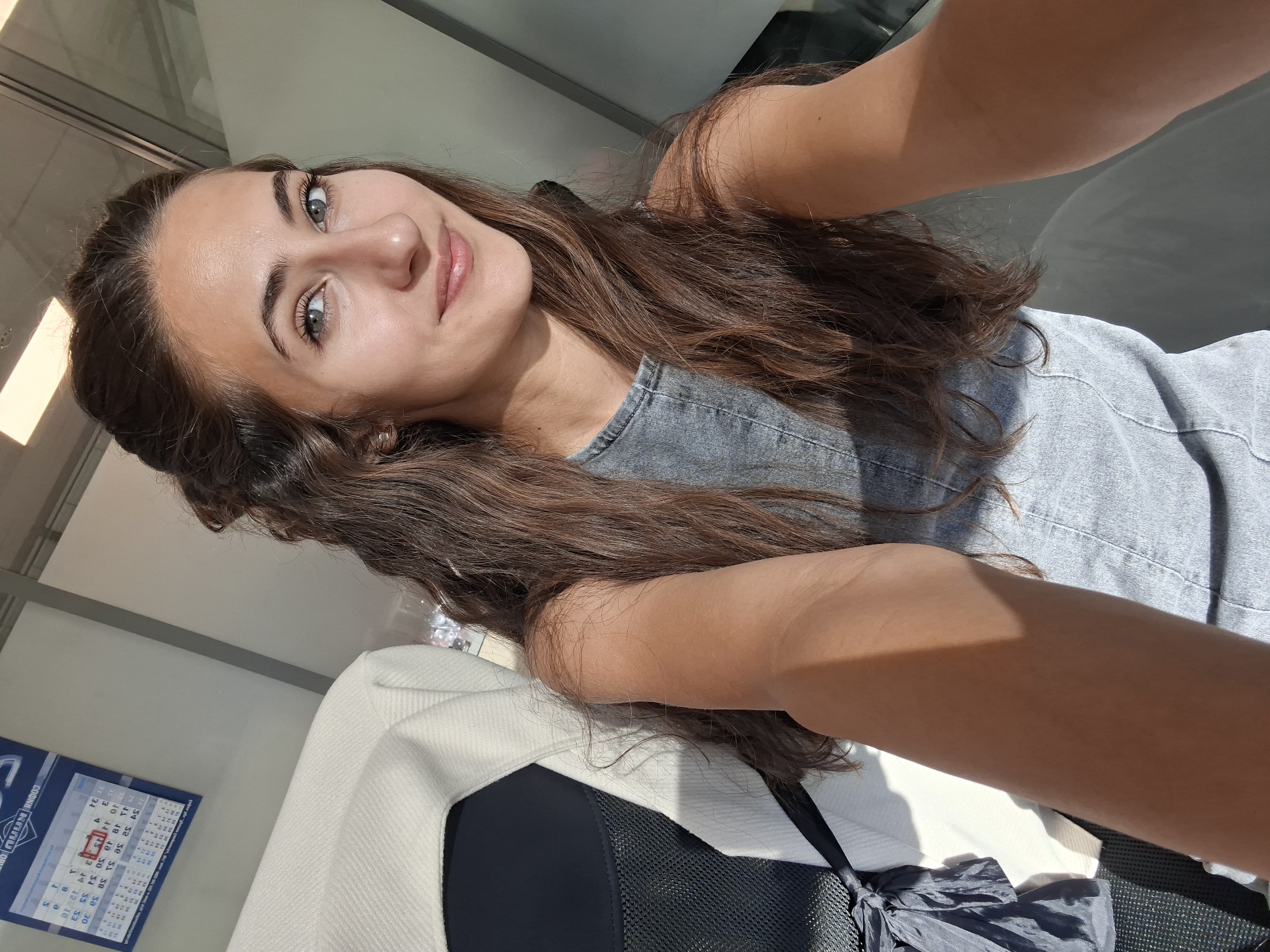 A young woman with long, wavy brown hair and green eyes smiles at the camera, taking a selfie. She is wearing a denim-like top and her arm is in the foreground, illuminated by sunlight. In the background, a white and dark-colored seat is visible, along with a calendar hanging on a wall. The environment appears to be indoors with natural light streaming in.