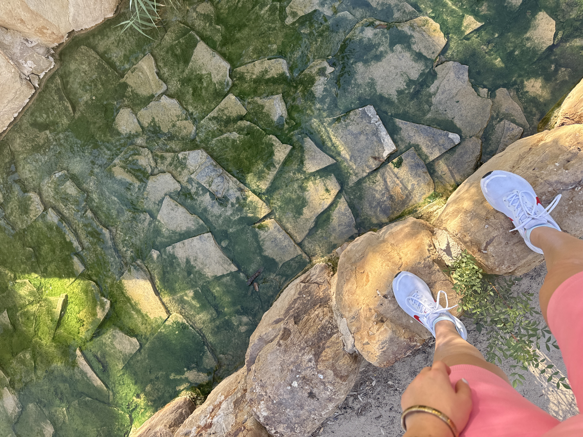 The image shows a top-down view of a person's legs and feet standing on large, rough rocks bordering a shallow, rocky stream. The stream bed is composed of irregularly shaped, flat stones covered in green algae. Sunlight casts dappled shadows on the water and rocks. The person is wearing pink shorts and white sneakers with red accents. One hand, adorned with a gold bracelet, rests on the rocks near their shorts. This scene is likely located in the Barranco de la Mula near the city of Murcia, Spain, a natural area known for its rocky terrain and occasional watercourses.