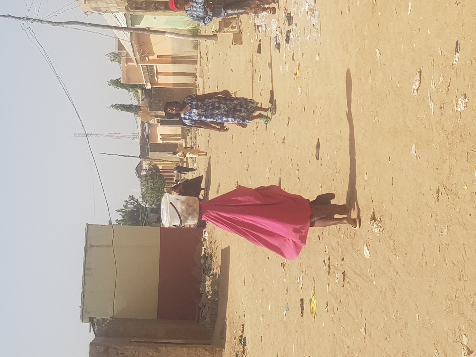 The image shows a street scene in Dabawa, Nigeria. The ground is dirt, and there is litter scattered about. In the foreground, a woman is walking away from the viewer, carrying a white bucket on her head. She is wearing a pink dress. In the background, another woman is walking, wearing a blue patterned dress. There are buildings on either side of the street. A few people are visible in the far background. There are utility poles and wires visible in the sky.