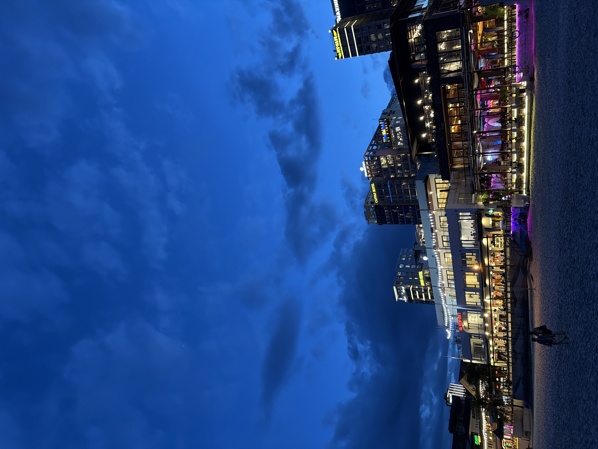 An outdoor, eye-level, wide shot captures a view of a beachside promenade at dusk in Đà Nẵng, Vietnam. The sky is a deep, rich blue, with a few wisps of dark clouds scattered across it. In the background, the city skyline is illuminated by artificial lights. Several multi-story buildings are visible, some with illuminated signage. One building prominently displays the words "CANVAS HOTEL" in white lettering. Another building has "OMAR HOTEL" lit in yellow. Further to the right, a tall building is lit with yellow vertical stripes and the word "MAXIM" is visible at the top.
In the middle ground, a strip of buildings lines the promenade, featuring shops and restaurants. One building with a white facade has "4D SEAFOOD" written across it. These establishments are brightly lit with warm, inviting lights, and many have outdoor seating areas with tables and chairs, some adorned with colorful purple and pink lighting. People are seated at some of the tables, and a few individuals are walking along the promenade.
The foreground is a sandy beach, appearing light and textured. Two figures, appearing to be a couple, are walking away from the camera across the sand towards the promenade. The overall scene depicts a lively evening atmosphere in a coastal city.