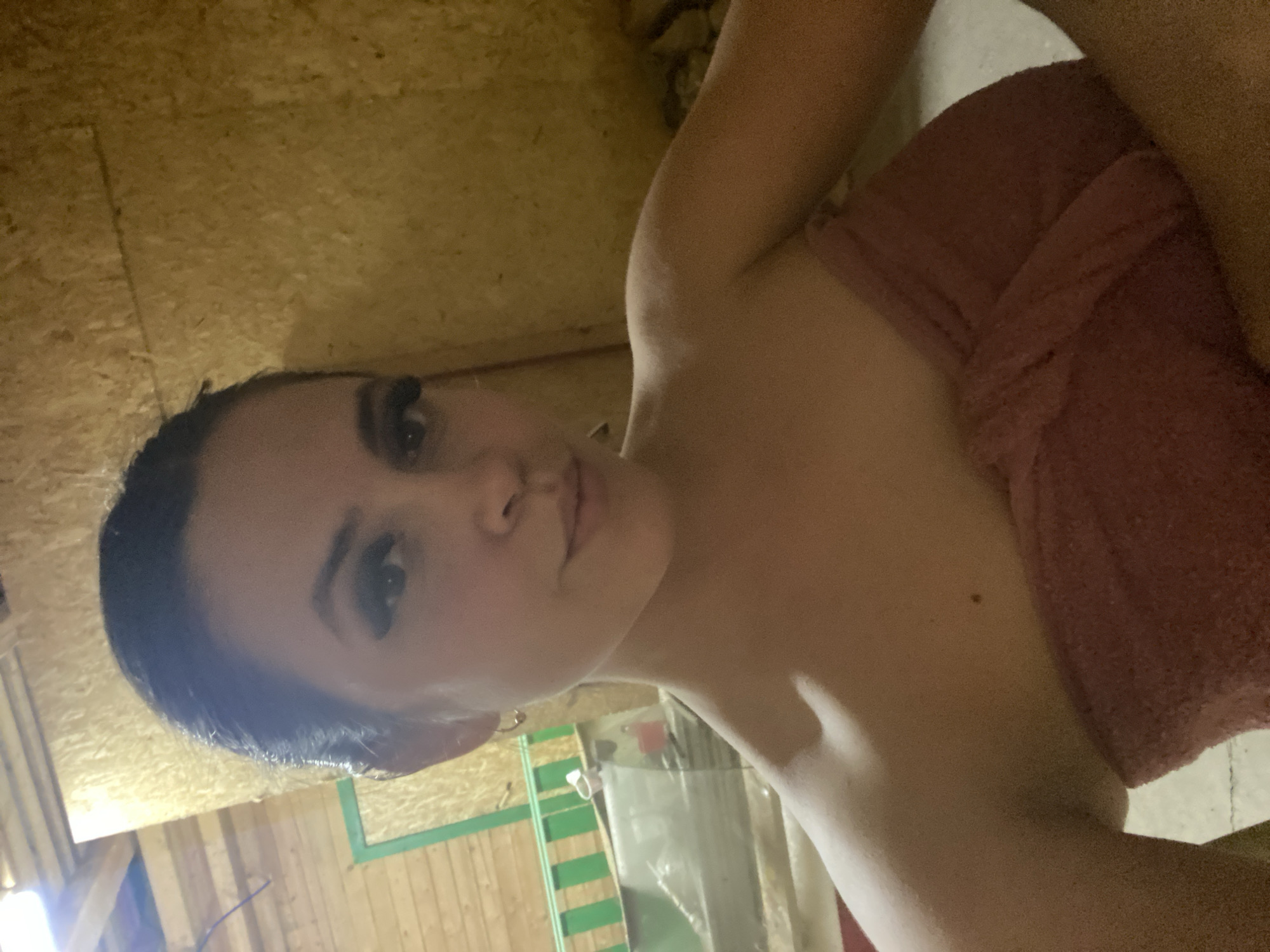 A person is partially visible, wrapped in a pink towel. The surrounding environment consists of light-colored wooden walls, potentially paneling. A blurry background suggests a possible interior scene with pipes and additional green and white elements. The location is unknown.