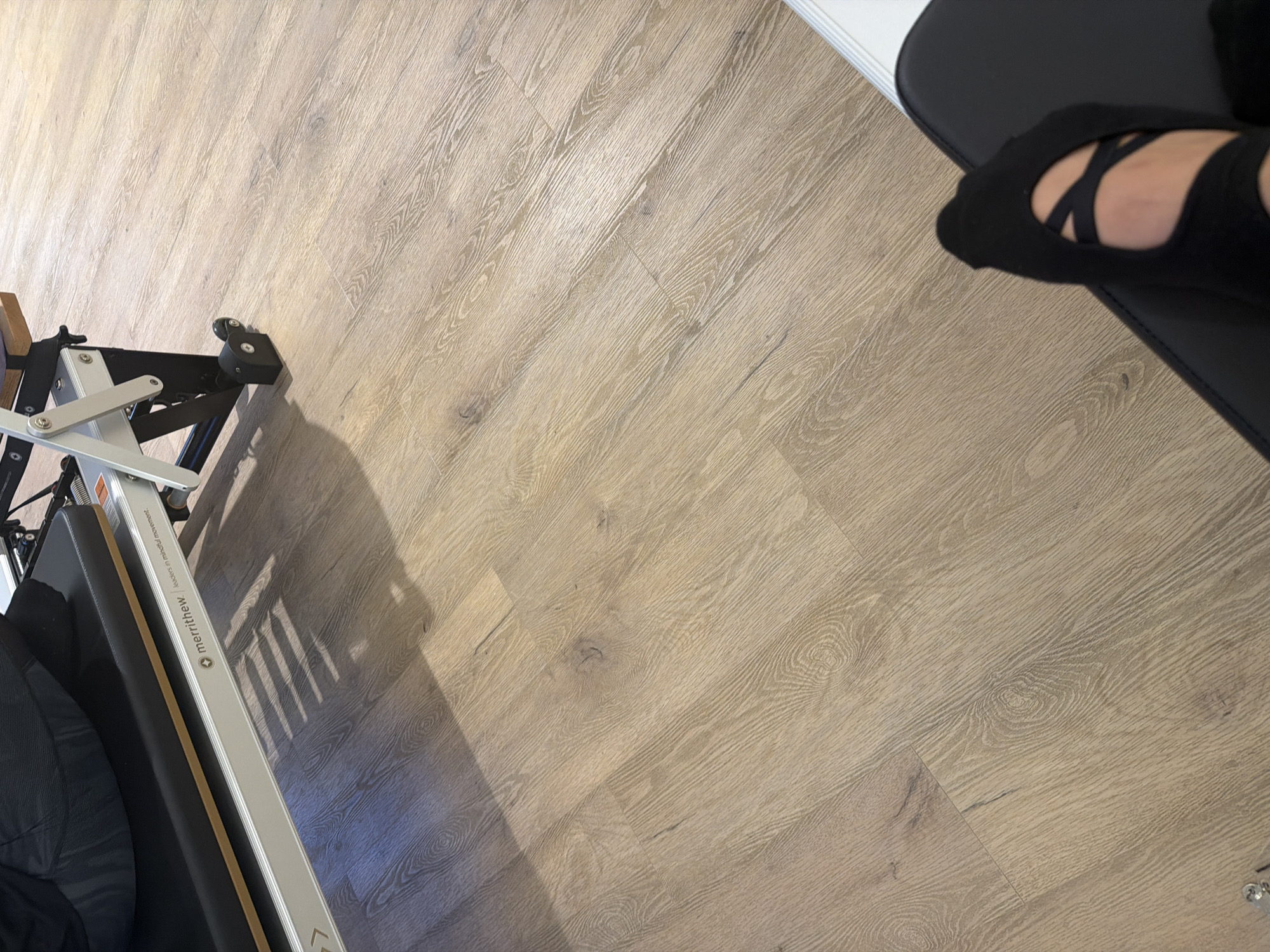 In a room with light wood flooring, a black padded surface of a pilates reformer is visible. The reformer has silver metal framing and a brand name "merrithew." A shadow from the reformer is cast across the floor. In the foreground, a person's feet are visible wearing black pilates socks with criss-cross straps. The room is likely a pilates studio in Boreti, Montenegro.