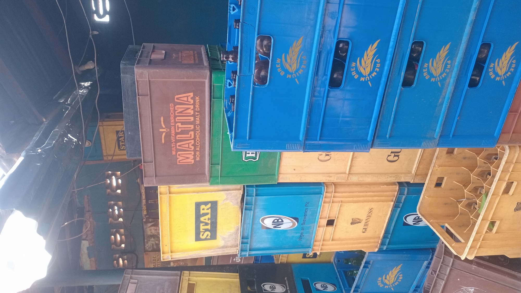 The image depicts an outdoor storage area, possibly a shop or market stall, in Jalingo, Nigeria. Stacks of plastic crates, many containing bottles, are the primary focus. Several crates are labeled with brand names, including "MALTINA," "STAR," "NB" (likely Niger Breweries Plc), and "GUINNESS." The crates are predominantly yellow, blue, green, and brown. Some bottles are visible within the crates, appearing to be beer or malt drinks. The background shows a corrugated metal roof supported by wooden beams, and a partially visible blue wall with barred windows. The overall impression is of a functional, perhaps slightly cluttered, storage space. There are no people visible in the image.