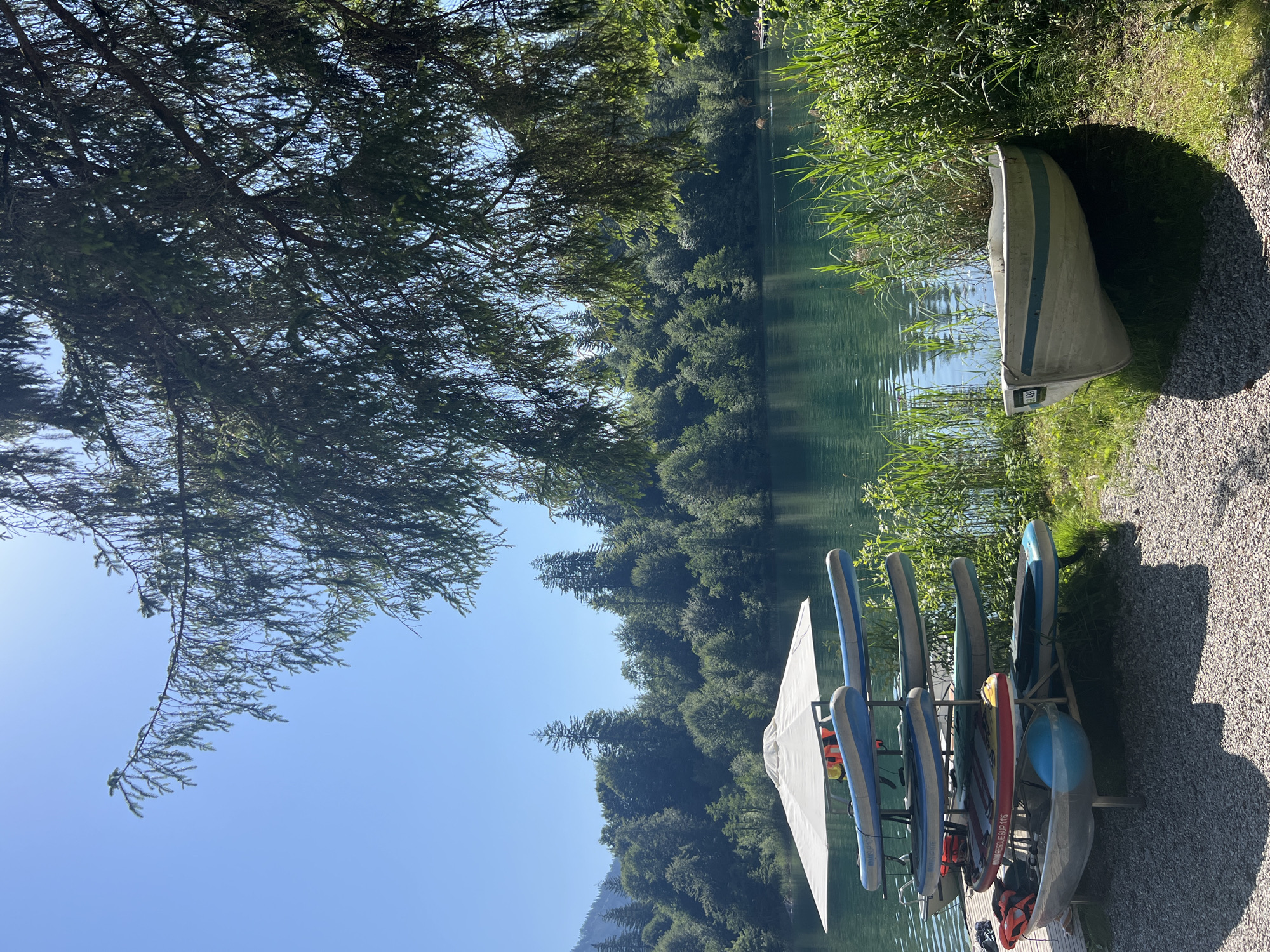 In an outdoor scene, a body of water is surrounded by lush green trees under a clear blue sky. In the foreground, several paddleboards are neatly stacked on a rack. To the right of the rack, a small white and blue rowboat rests on the grassy shore. A large white umbrella stands behind the paddleboard rack, providing shade. The scene appears to be a recreational area by a lake, likely in a natural or park setting, with no people visible.