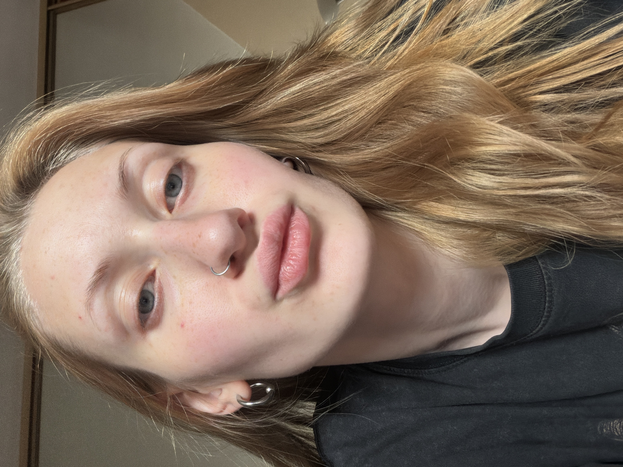 This is a close-up, low-angle, indoor, portrait photograph of a young person with fair skin and long, wavy, blonde hair. They are wearing a black t-shirt and silver hoop earrings in both ears, as well as a silver nose ring. The subject's face is partially obscured by their hair and the orientation of the photo. Their eyes are blue-grey and their lips are slightly parted. The background is blurred and appears to be a room with neutral-toned walls and a hint of furniture on the left side, possibly a closet or a mirror reflecting part of the room. The lighting suggests it is daytime, with natural light illuminating the subject's face. No text is visible in the image. The overall mood is casual and relaxed.