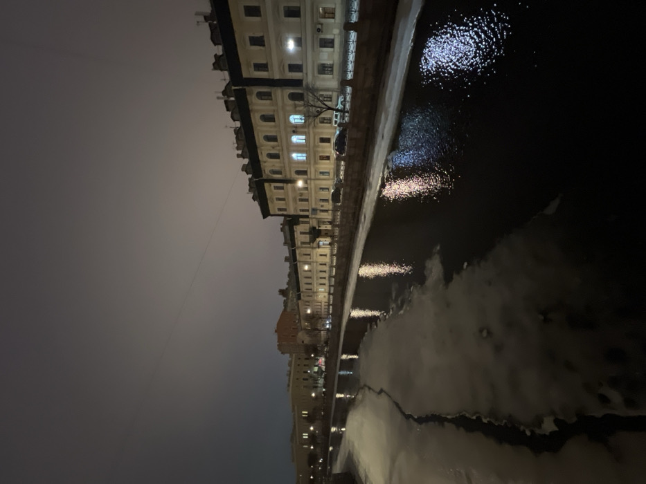 The image captures a nighttime, elevated view of a partially frozen canal in Saint Petersburg, Russia, identifiable as the Griboyedov Canal. The dark, overcast sky dominates the upper portion of the frame. The canal's surface features large, irregular patches of light-colored ice, particularly prominent on the left, contrasting with sections of open, dark water. Artificial lights from the embankment and buildings reflect brightly on the water, appearing as shimmering vertical streaks of white and yellowish-orange.
Along the right bank, a stone embankment with a decorative railing runs parallel to the canal. Behind this, a street is visible, populated by several parked vehicles, including dark cars and a white van. A continuous row of multi-story, historic-style buildings with light-colored facades lines the street. These structures feature numerous windows, some of which are illuminated from within, casting warm light; one second-story window prominently displays a blue-white light. Dark rooftops with visible chimneys crown the buildings. A bare deciduous tree stands between the street and the buildings. Further down the canal on the left bank, additional buildings with illuminated windows are discernible in the distance. No individuals are visible in the scene.