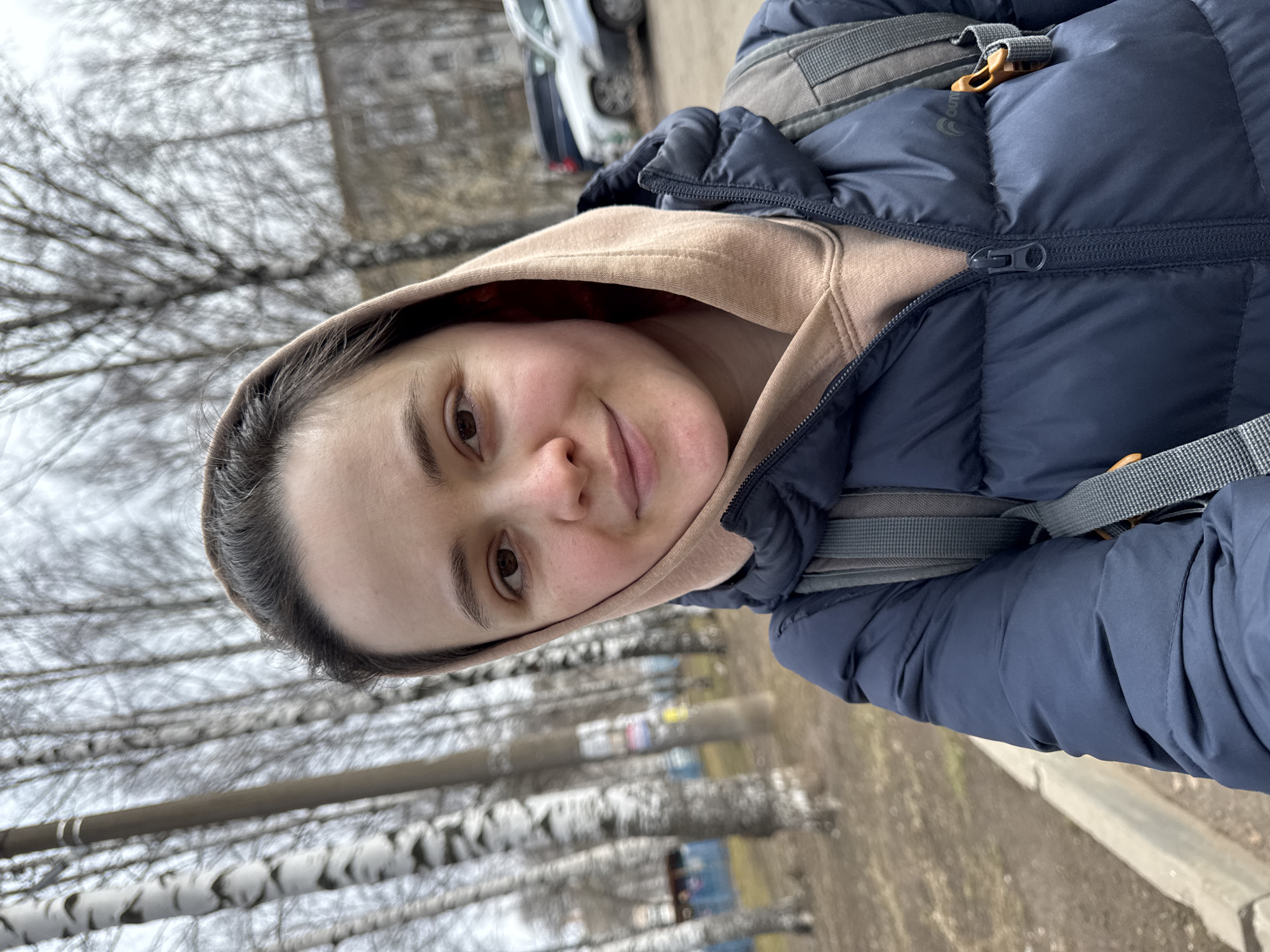 A young woman, likely in her late teens or twenties, is featured in a close-up, outdoor portrait. She has dark hair, dark brown eyes, and a pleasant, gentle smile. Her head is slightly tilted, and she looks directly at the camera. She is dressed in a light beige hooded sweatshirt worn under a dark blue puffy jacket. The straps of a gray backpack are visible on her shoulders.
The setting appears to be an urban or suburban outdoor area during the daytime. The trees in the background are bare, with light-colored bark reminiscent of birch trees, suggesting a colder season like late autumn, winter, or early spring. The sky is overcast, creating a soft, diffused light. In the blurry background, a paved path or road, some indistinct buildings, and a car can be seen.
The woman's expression conveys a relaxed and amiable mood. On the upper left chest area of her blue jacket, a small, circular, light-colored logo is visible, but the text or emblem within it is too indistinct to be clearly read. The overall impression is a casual, everyday moment captured in a cool climate, consistent with the Izhevsk, Russia location context.