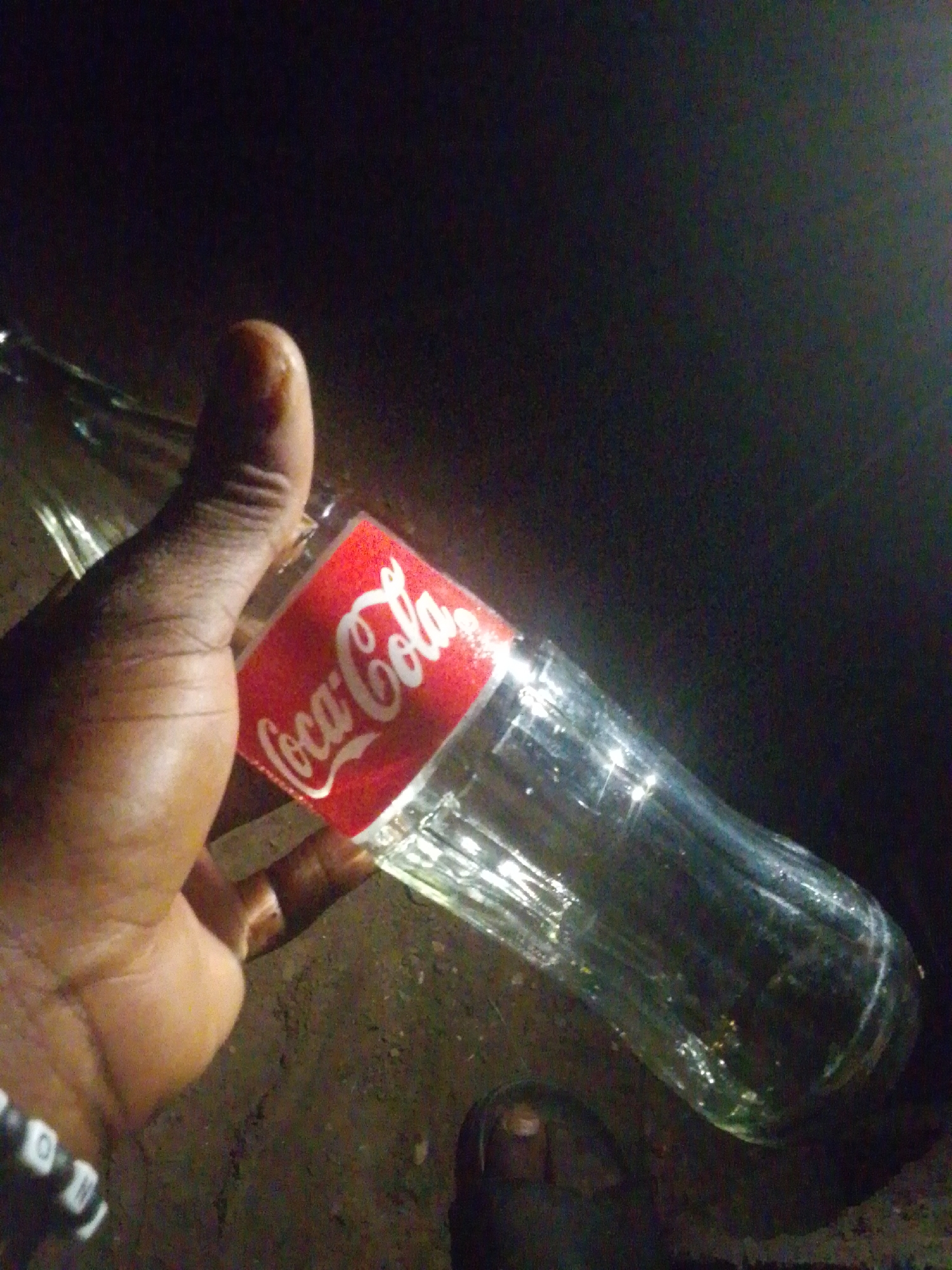 The scene is set in an unknown location in the city of Unknown, Unknown. The image displays a human hand with dark skin holding an empty, clear glass Coca-Cola bottle. The hand's thumb is visible on the upper part of the bottle's body, with fingers wrapped around it. A bracelet featuring black and white cylindrical beads is worn on the wrist. The glass bottle has a red label with the white "Coca-Cola" logo. The background is dark and indistinct, indicative of low light conditions. The ground surface beneath the bottle and a partially visible human foot, clad in a dark open-toed sandal, appears to be rough, dark earth or pavement. Reflections are present on the glass surface of the bottle due to an unseen light source.