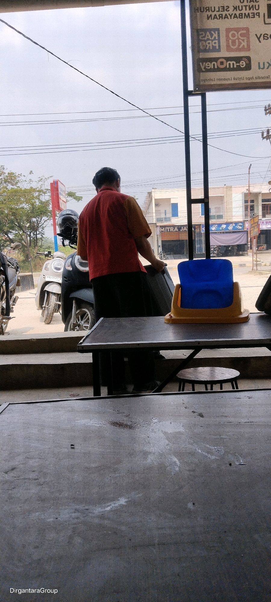 The image depicts an outdoor scene, likely in Sirih Sekapur Perkembangan, Indonesia. A man wearing a red and yellow shirt and black pants is seen from the back, holding a dark-colored object. He is standing in front of a scooter. In the background, buildings are visible, and there are power lines overhead. Two tables are in the foreground, one with a small, round stool underneath it. A blue and yellow object, possibly a container, sits on the table. The lighting suggests it is daytime.