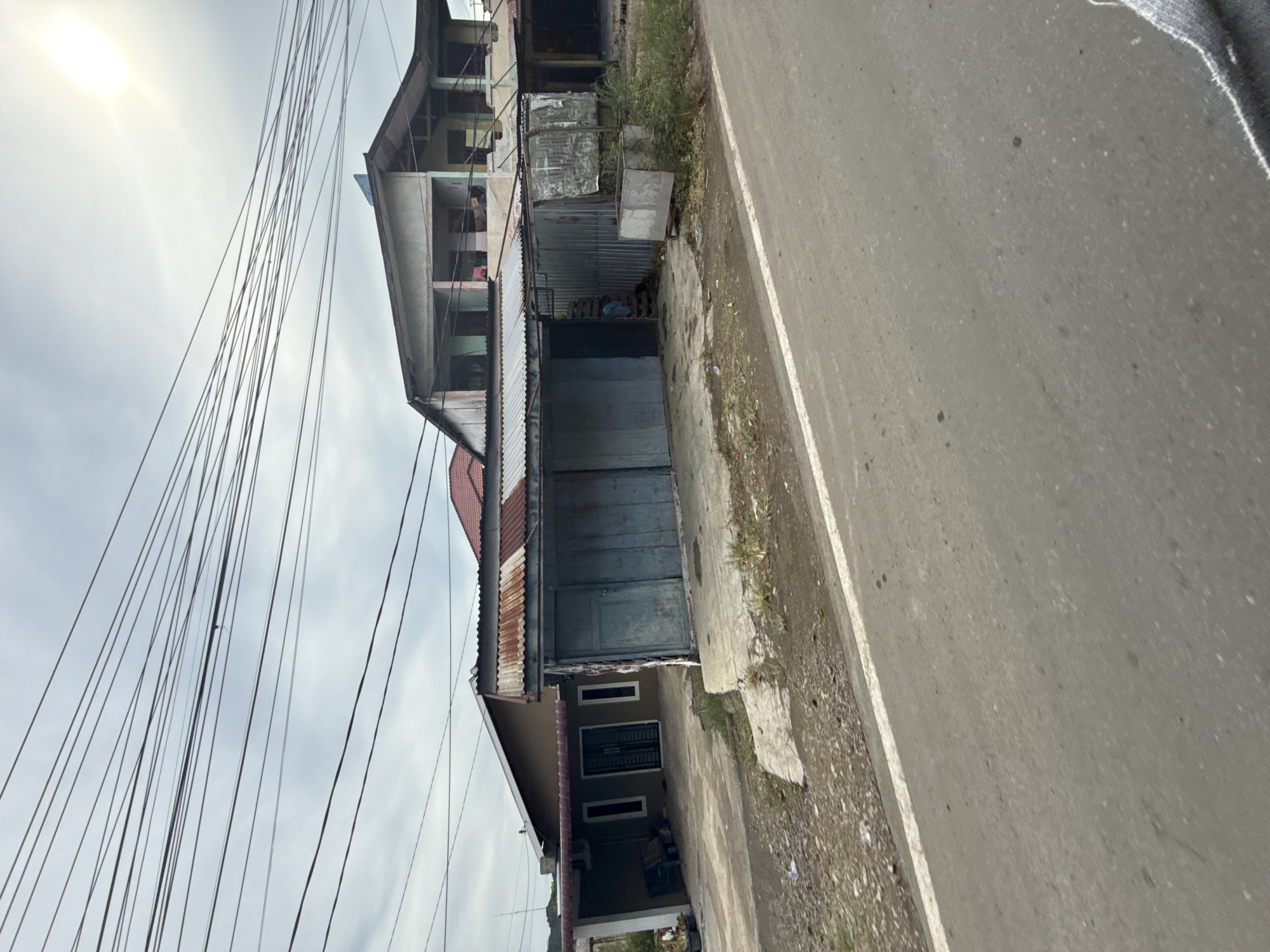 The image depicts a roadside scene in Banda Aceh, Indonesia, under an overcast sky with visible power lines crisscrossing overhead. In the foreground, a paved road with a white line marking its edge runs diagonally from bottom left to top right. Adjacent to the road, a gravelly area with some patches of dry grass and debris is visible.
The main focus of the image is a row of buildings. On the left, a single-story building with a brown exterior and a covered porch area is partially visible. The porch has a concrete floor and two white-framed windows and a door with a metal grille are visible on the building's facade. A blue bench sits under the porch.
To the right of this building is a more dilapidated structure with a corrugated metal roof, showing signs of rust. This section appears to be a shop or storefront with two large, weathered, light blue sliding doors. Next to this, a corrugated metal fence encloses a small area with a step leading up to a doorway. Further to the right, a large, rectangular, metal bin or container sits on a frame, surrounded by overgrown green vegetation.
Behind these lower structures, a multi-story building is visible. The upper floors appear to be constructed with a lighter colored material, possibly concrete or stucco, and have open window frames and balconies. The overall impression is one of a working-class or older commercial area, possibly with residences above the shops. The presence of extensive power lines suggests a typical urban or semi-urban environment.