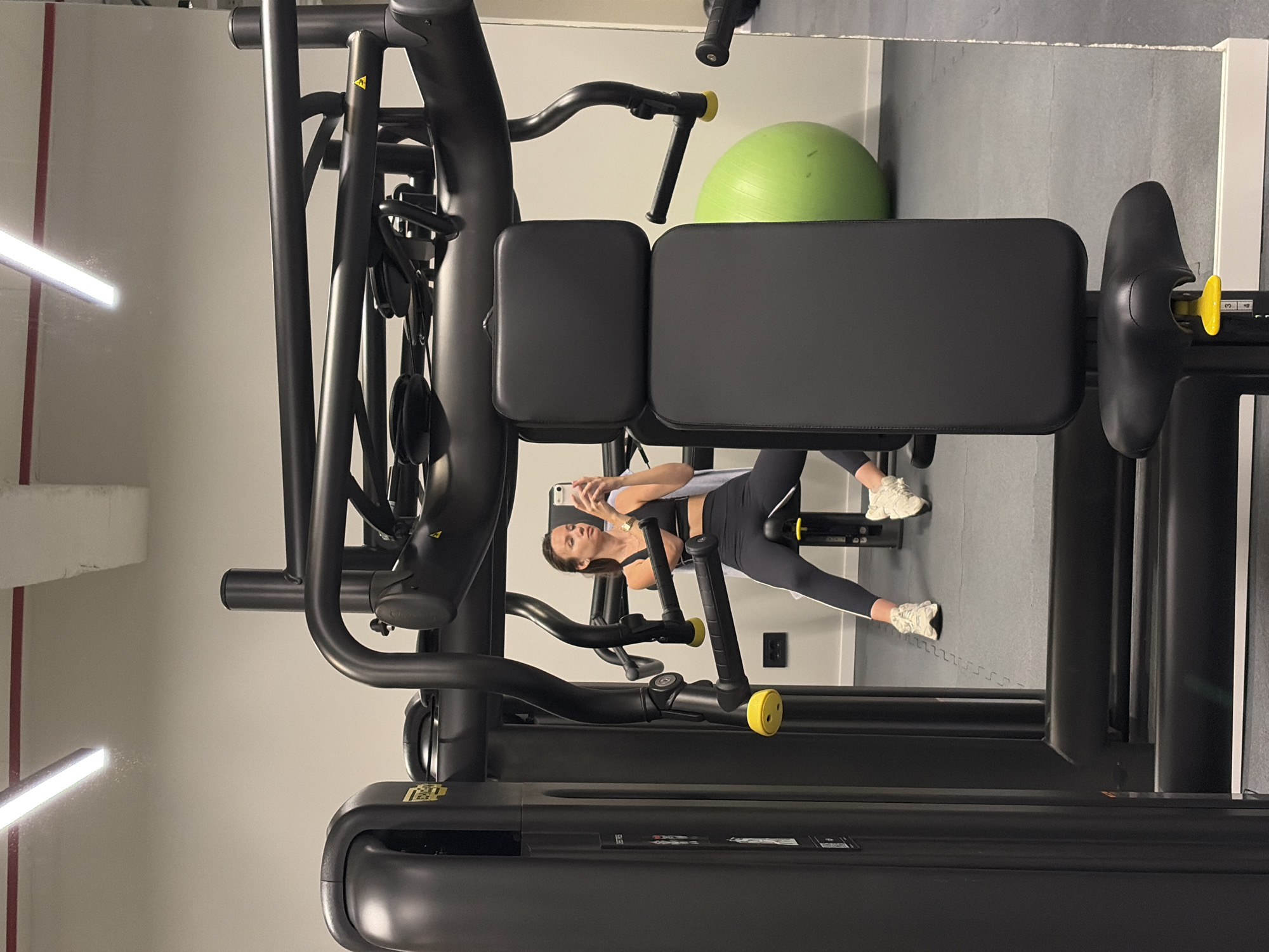 A gym scene at an Unknown location within Unknown, shows a woman seated on a black exercise machine, possibly engaging with a smartphone. The machine has various metallic and cushioned components. Behind her, there is a large green exercise ball, and the flooring appears to be made of interlocking gray mats. The gym’s interior is lit with linear light fixtures, casting light across the off-white and gray walls.