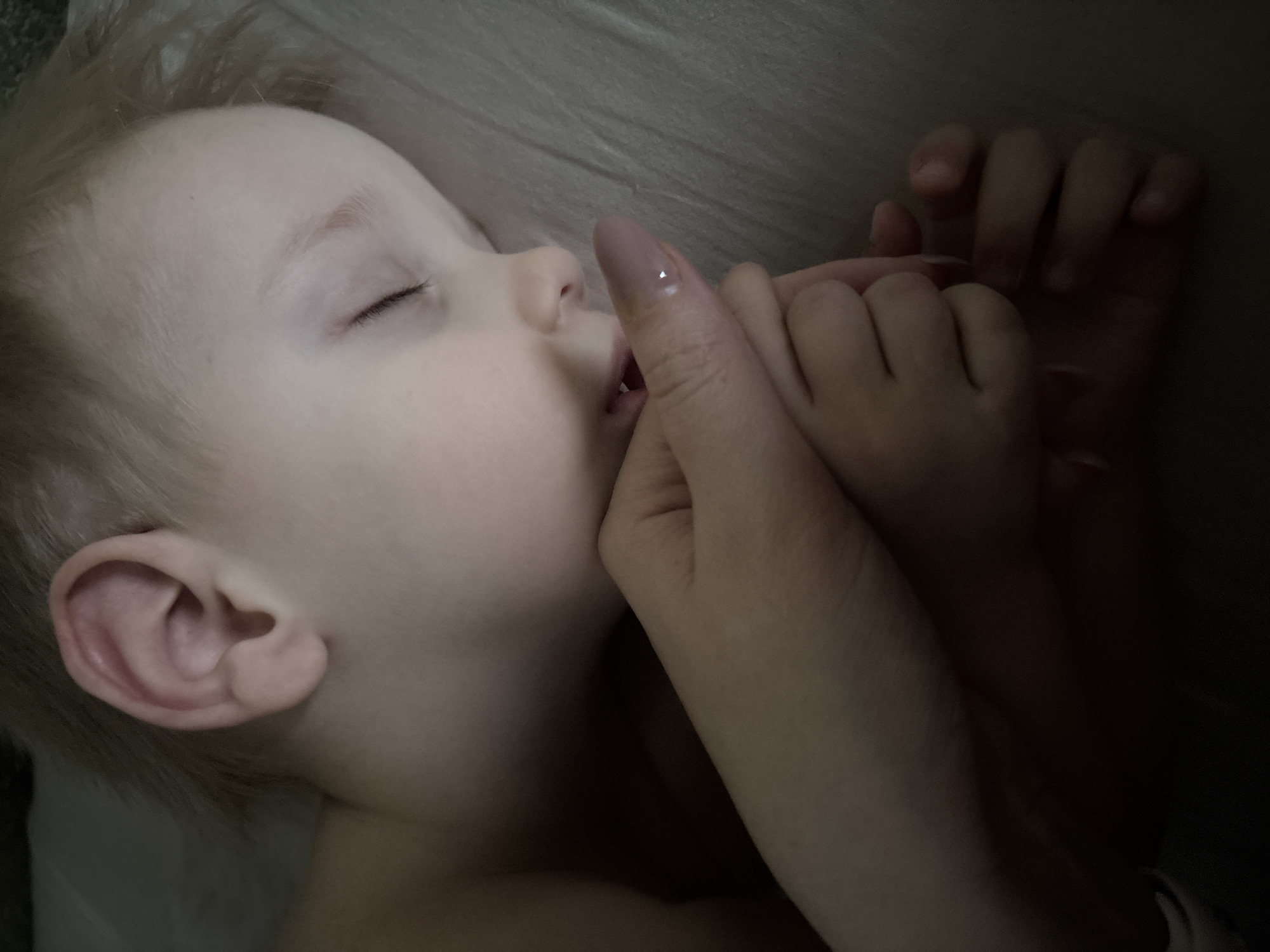A fair-skinned, blond toddler is sleeping in a dimly lit environment. The child's eyes are closed, and a partially visible adult hand is holding the child's hand, with one finger near the toddler's mouth. The background appears to be a light-colored sheet or fabric. There is no information available to establish the location as Chaikovsky, Russia.