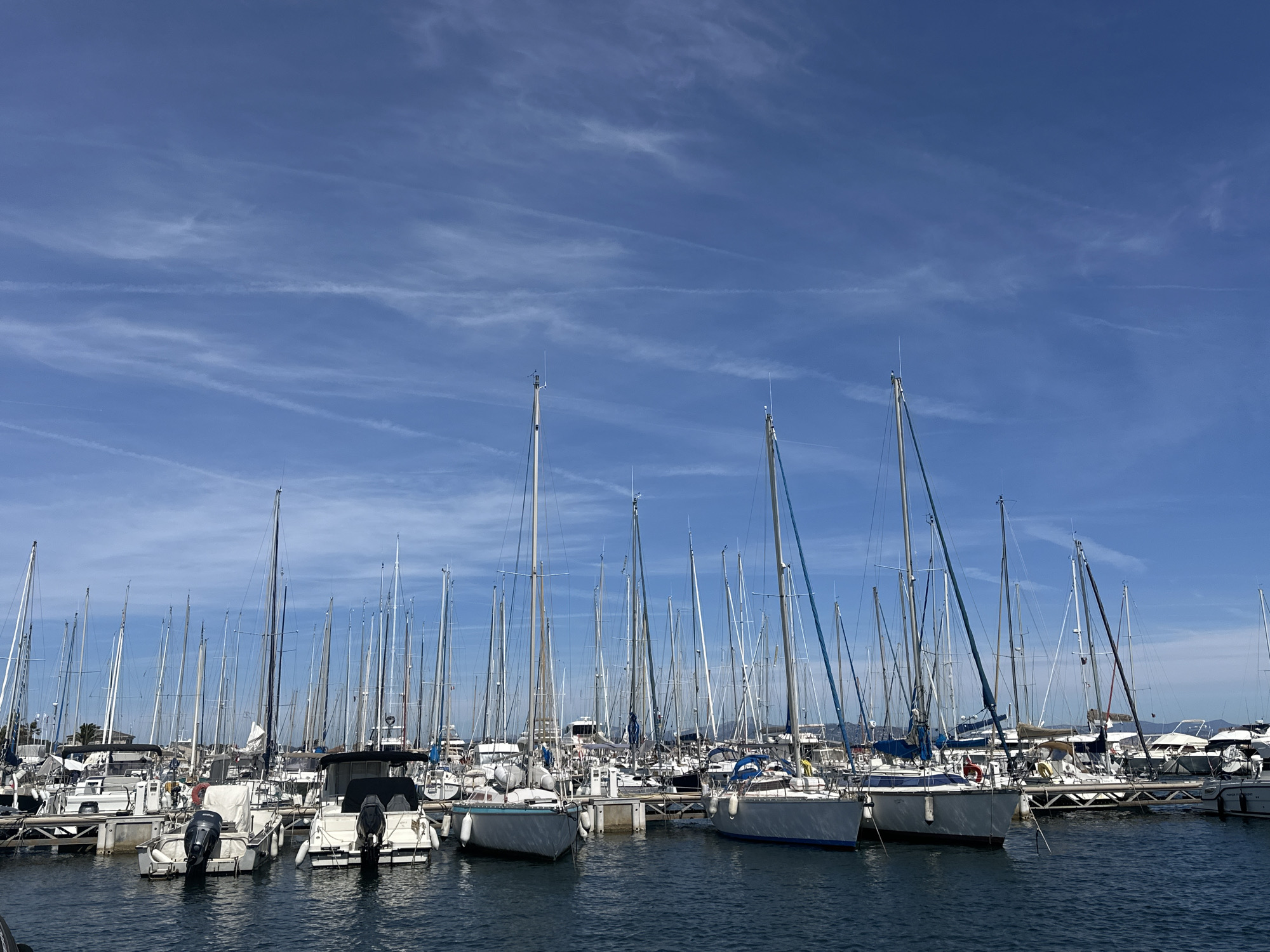 This image depicts a crowded marina filled with numerous sailboats docked in calm blue water under a bright sky. The sky is a vibrant blue with thin, wispy clouds and contrails. The marina is packed with sailboats of various sizes, their masts forming a dense forest against the sky. Many of the boats are white with dark blue or grey accents. Some of the boats have outboard motors visible at the stern. The boats are moored to wooden docks. The background shows a distant hazy landscape with some structures, possibly indicating the city of Hyères, France. The scene suggests a clear, sunny day. No people are visible in the image.