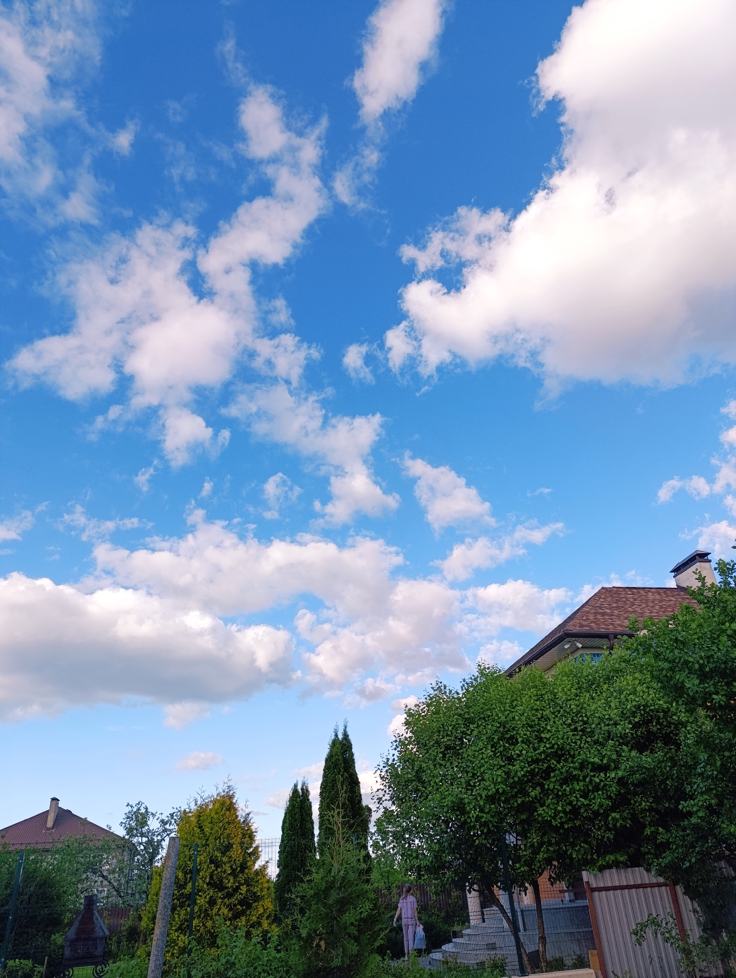 The image shows a residential scene under a bright blue sky dotted with fluffy white clouds. A two-story house with a brown tiled roof is partially visible on the right side, framed by lush green trees. In the lower left, a glimpse of another house with a red roof and some greenery is seen behind a fence. Several coniferous trees are positioned centrally. A person wearing light-colored clothing can be seen walking on steps, holding a bag or container. A metal fence runs along the right side of the image.