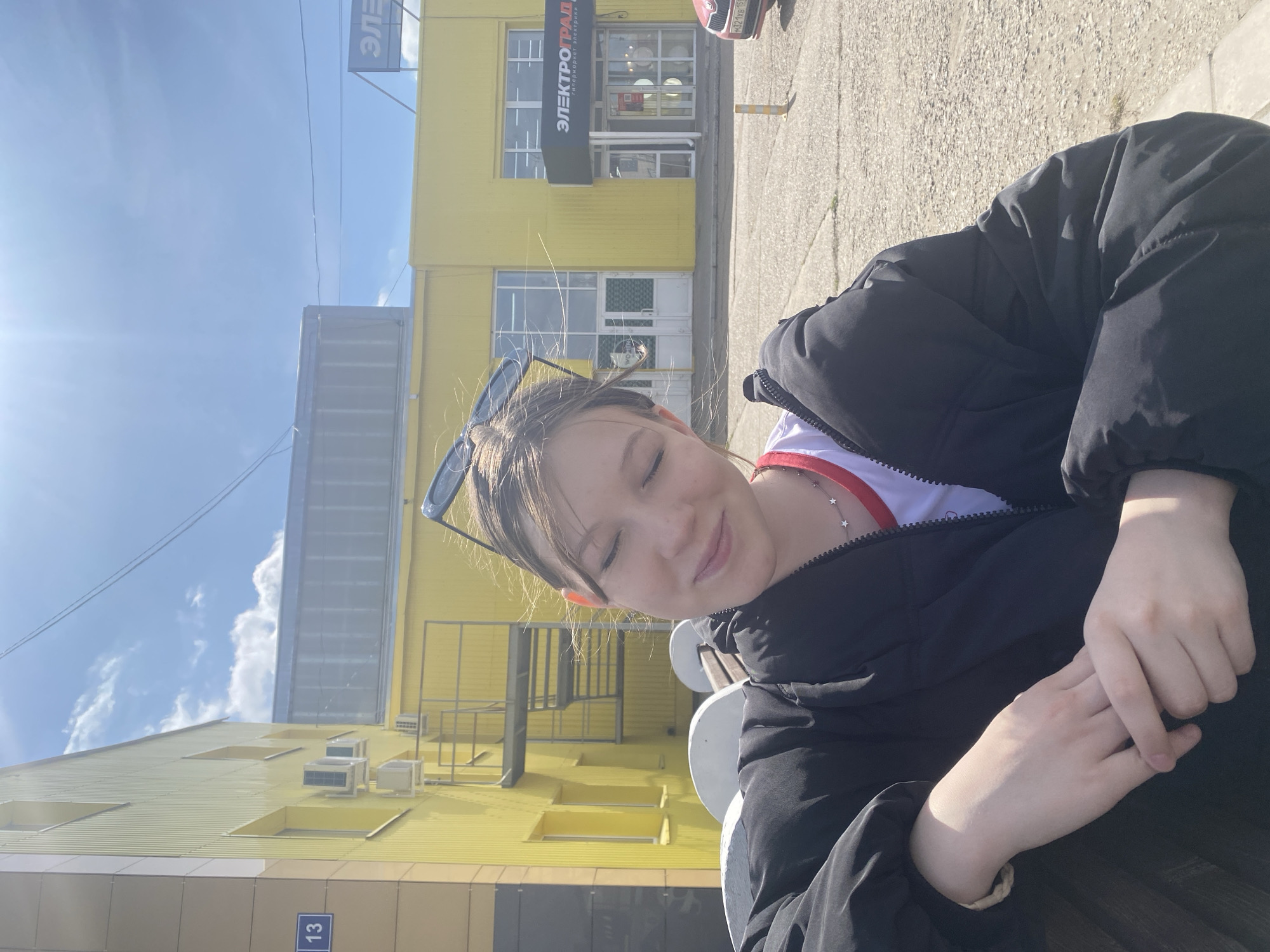 The image captures a serene outdoor scene, featuring a young person in the foreground against the backdrop of a bright yellow commercial building under a clear sky.
The subject, appearing to be a young individual with fair skin and light brown hair, occupies the right side of the frame. They wear sunglasses perched on their head and are dressed in a dark, possibly black, puffer jacket over a white and red garment. Their eyes are gently closed, and a subtle smile plays on their lips, conveying a sense of peace or contentment. Their hands are clasped together, resting in the lower right portion of the image.
In the background, a striking, multi-story yellow building dominates the scene. It features corrugated metal siding and numerous windows, some reflecting the blue sky. Grey metallic external stairs or railings are visible on the building's facade. On the roof, several air conditioning units are installed. The ground beneath is paved with asphalt or concrete. A small blue sign with the number "13" is visible near the base of the building, and a partial view of a red car with a visible license plate is seen near the building's entrance.
The sky is a bright, clear blue with scattered white clouds, indicating sunny weather. The strong light and visible shadows suggest the photograph was taken during midday or early afternoon.
Visible text on the yellow building's facade reads "ЭЛЕКТРОГРАД приборы электрики интерьеры", which translates to "ELECTROGRAD appliances electrical interiors". A partial sign displaying "ЭЛЕ" is also visible in the upper left. The license plate on the red car is "H321YB 159", where "159" is the regional code for Perm Krai, confirming the location as Chaikovsky, Russia.