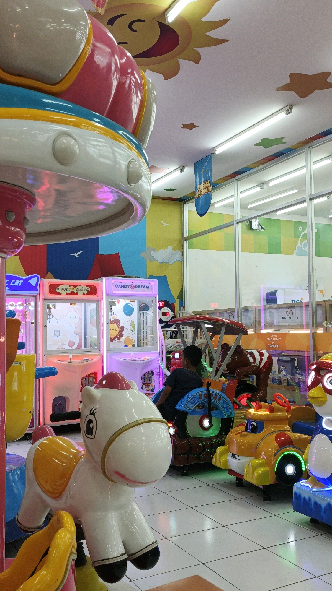 This is a vertical, eye-level photograph taken indoors, likely in an amusement arcade or children's play area. The scene is brightly lit by fluorescent lights on the ceiling, some of which are visible. The ceiling itself is decorated with a cartoon sun and several colorful star shapes.
In the foreground, a white toy horse with a pink mane and red hooves is the primary subject, angled towards the viewer. Its eyes are wide and black, and it has a pink smile. Behind the horse, a young boy with dark hair and wearing a dark blue t-shirt is seated on a rideable toy that resembles a dark brown horse with red and white striped reins. The boy is looking to his right.
To the left of the horse and the boy, there are several brightly colored arcade machines. One machine is pink and white with the text "CANDY DREAM" in pink letters. Another machine, partially visible, is pink and has a yellow dispenser. To the far left, a yellow object, possibly a part of another ride, is visible.
Behind the boy and the arcade machines, there is a wall decorated with a colorful mural featuring red tents, blue mountains, white clouds, and a yellow sky with white birds. Beyond this wall, through a glass partition, there are shelves with various items, including what appear to be drink bottles and boxes.
To the right of the boy's ride, there are other colorful children's rides. A yellow car-shaped ride with blue and red accents and green lights is prominent. Next to it is a blue ride with a white and red face, possibly a bird or cartoon character.
The floor is tiled with white square tiles. The overall atmosphere is cheerful and geared towards children's entertainment. The presence of arcade machines, rideable toys, and bright decorations suggests an indoor amusement venue. The mention of Desa Karangbenda, Indonesia, suggests this location is a local attraction within that area.